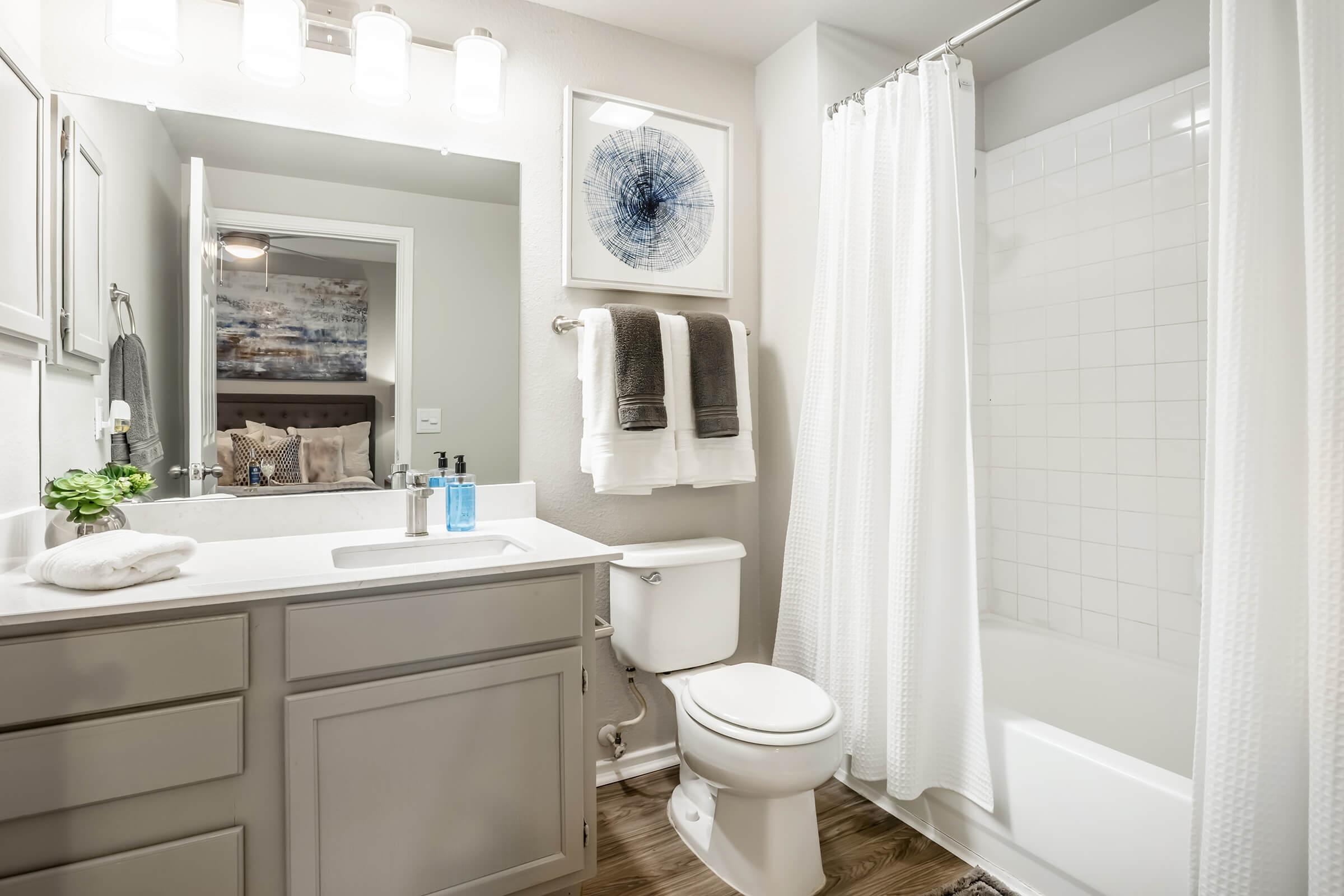 Modern bathroom featuring a white shower curtain, grey cabinetry, and a large mirror. There are neatly arranged towels on a rack and a small plant on the counter. A bathtub is visible alongside a toilet, with soft lighting creating a warm atmosphere.