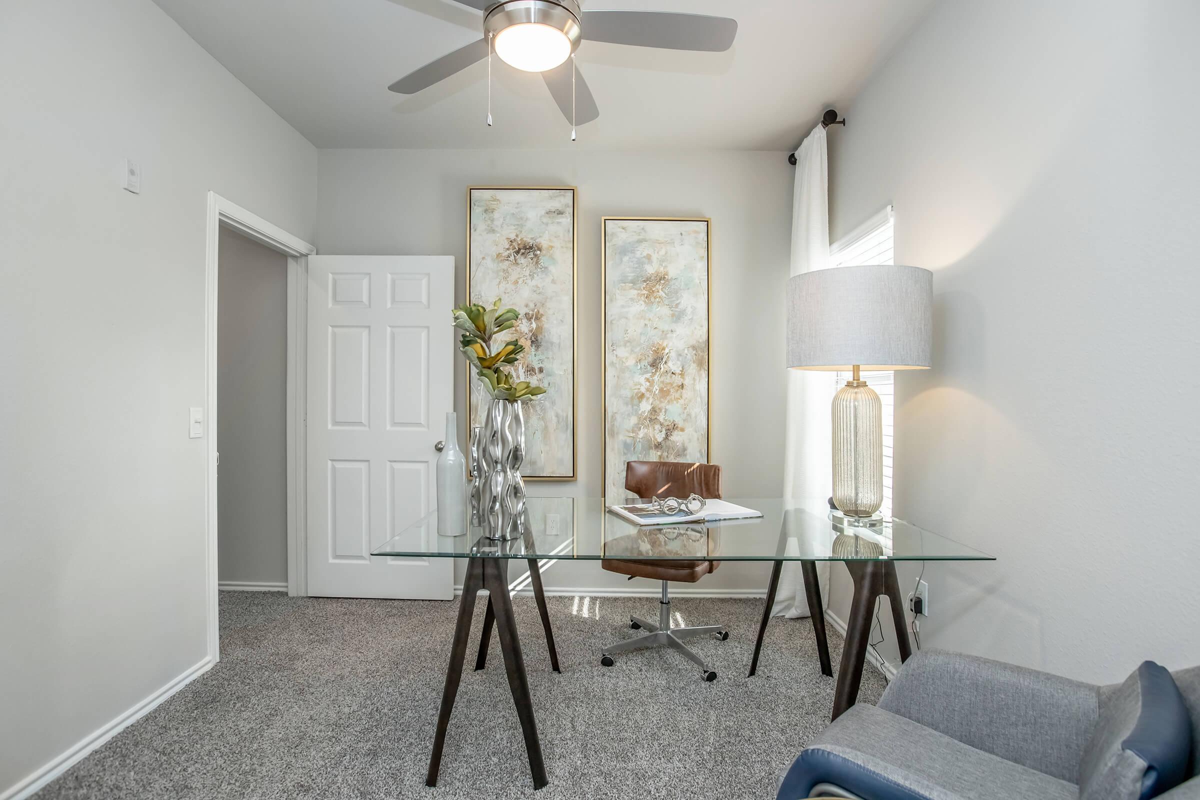 A modern home office featuring a glass desk with a notebook and decorative items, a stylish brown chair, a floor lamp with a large shade, and two abstract art pieces on the wall. The room has light gray walls, a ceiling fan, and a door leading to another area. Plush carpet covers the floor.