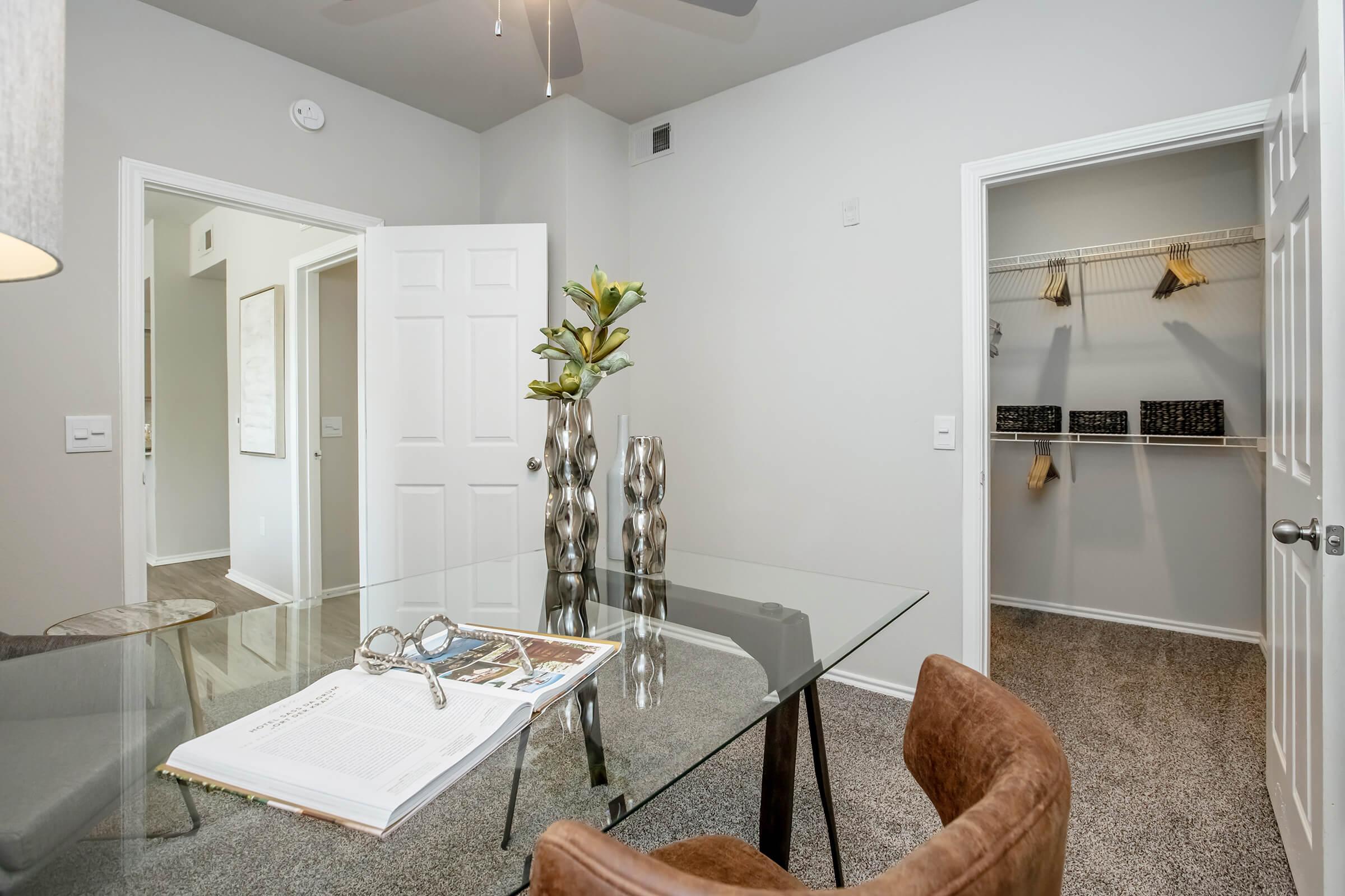 A modern interior office space featuring a glass desk with a book and decorative items. There is a stylish chair, a potted plant, and a closet with shelves in the background. The room has light-colored walls and a ceiling fan, creating a bright and airy atmosphere.