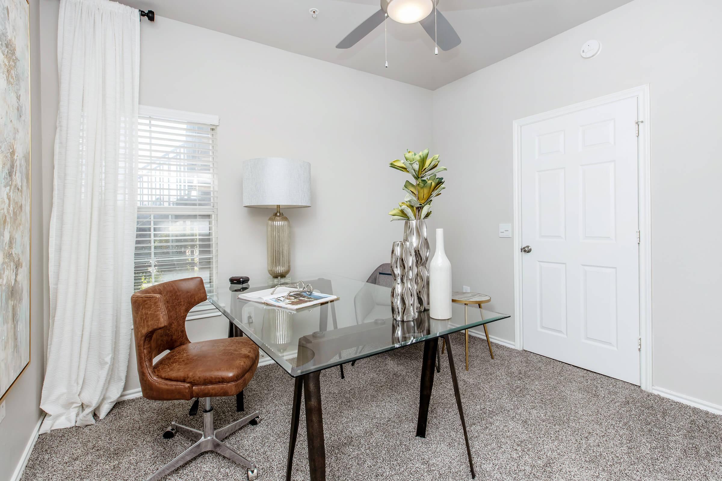 A modern office space featuring a glass-top desk with a decorative magazine, a stylish brown chair, a tall lamp, and vases with faux greenery. Light-colored walls and a window with white curtains create a bright ambiance. A closed white door is visible in the background.