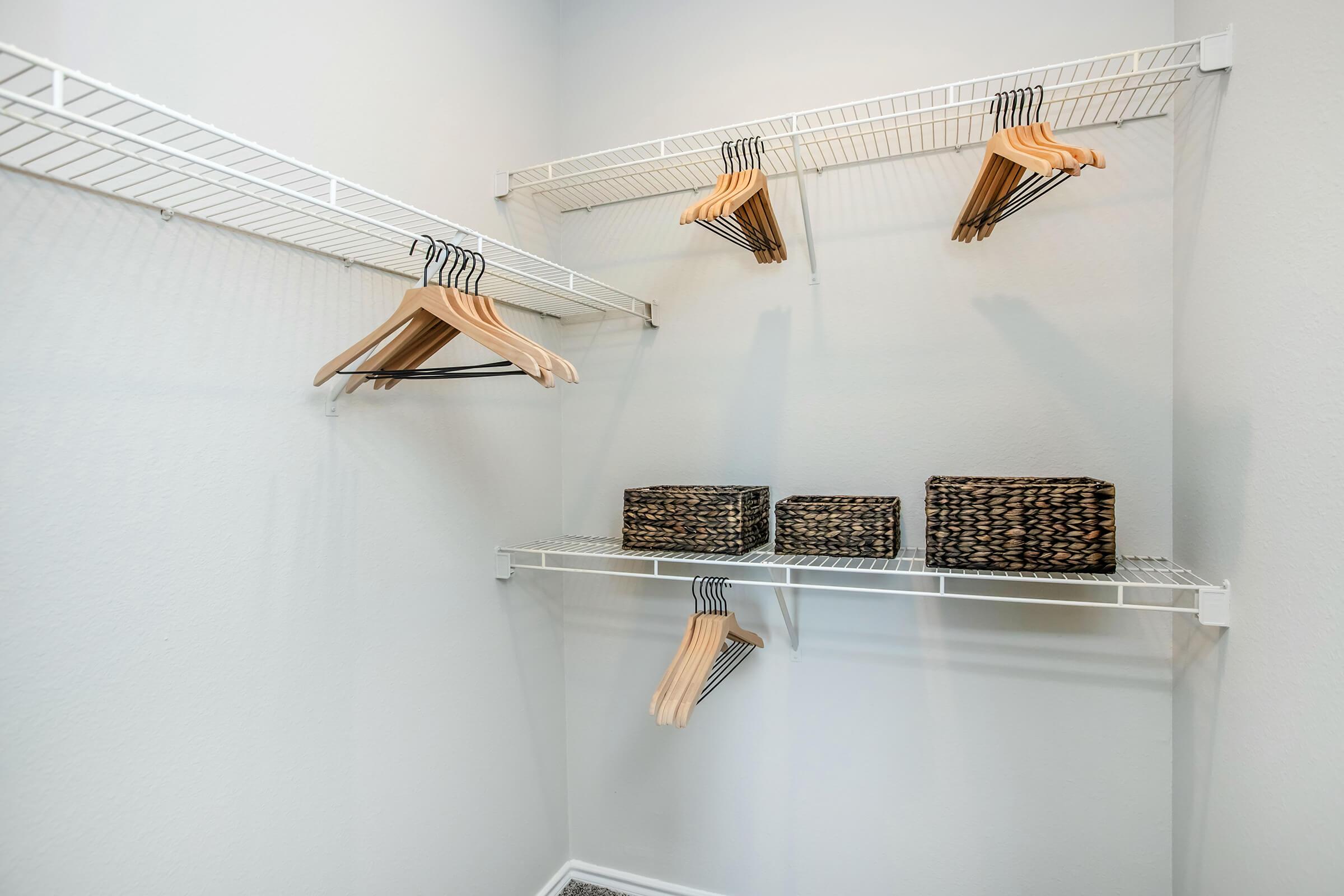 A clean and organized closet with white wire shelves. Several wooden hangers are hanging on the top shelf, while two wicker storage baskets are placed neatly on the bottom shelf. The walls are painted in a soft, light color, creating a spacious and tidy appearance.