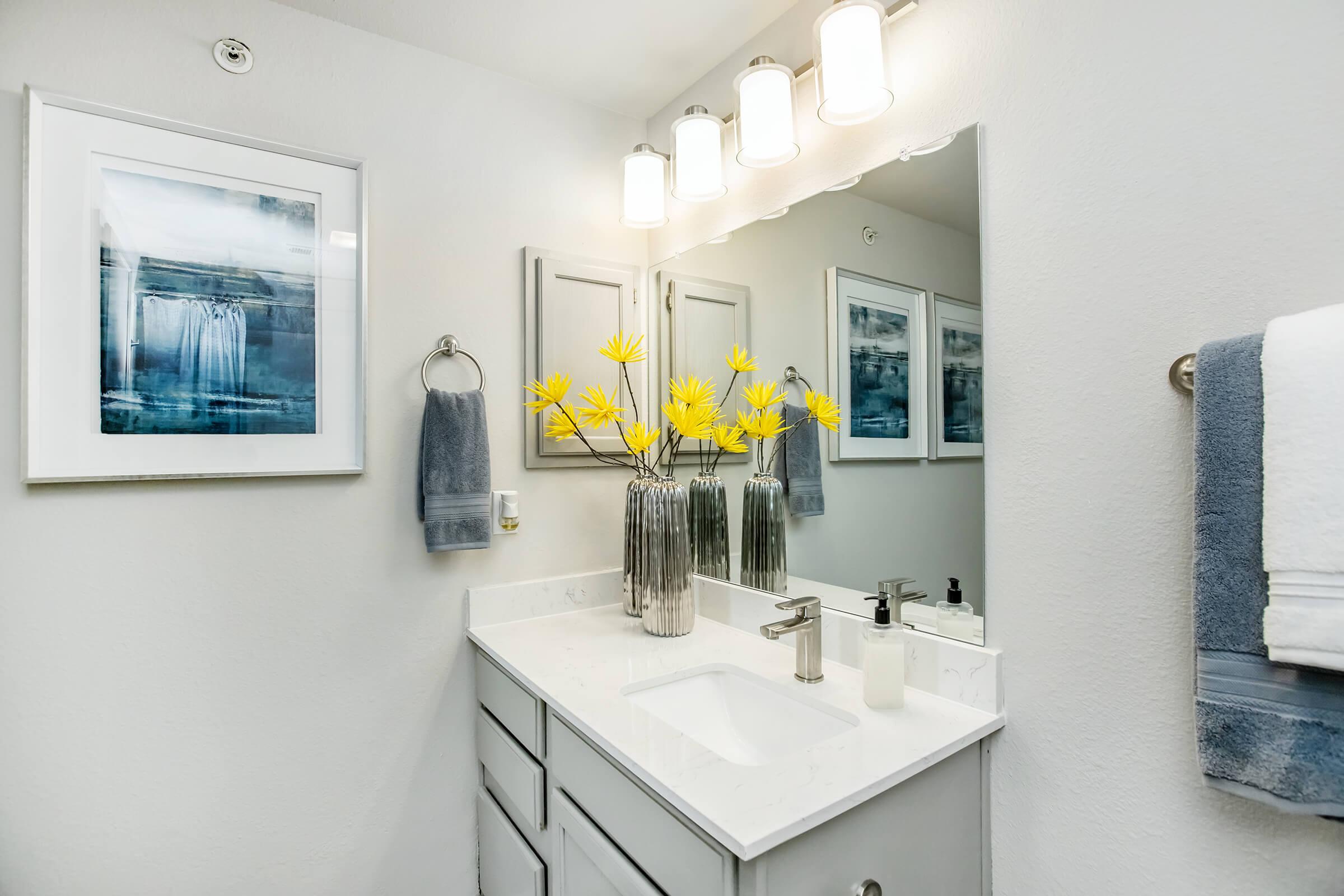A modern bathroom featuring a white vanity with a sink, a large mirror above, decorative towels, and a wall-mounted light fixture. A framed artwork of a waterfall hangs on the wall, and a vase with yellow flowers adds a pop of color to the space. The walls are painted in a light gray tone.
