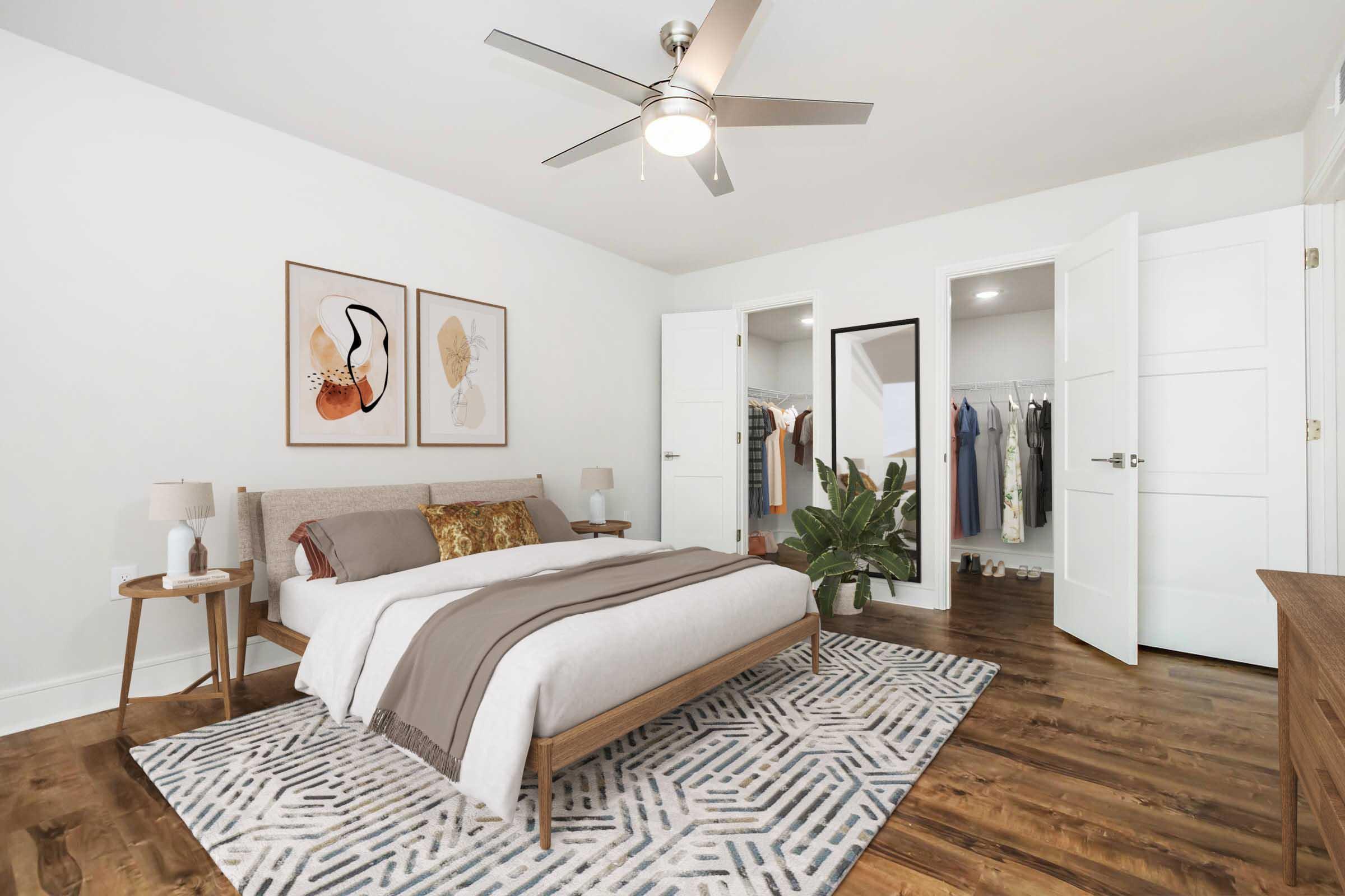 A modern bedroom with a light-colored wall and hardwood floor. The room features a bed with decorative pillows, two bedside tables with lamps, and framed artwork on the wall. A mirror and closet with hanging clothes are visible in the background, along with a potted plant. A ceiling fan is installed above the bed.