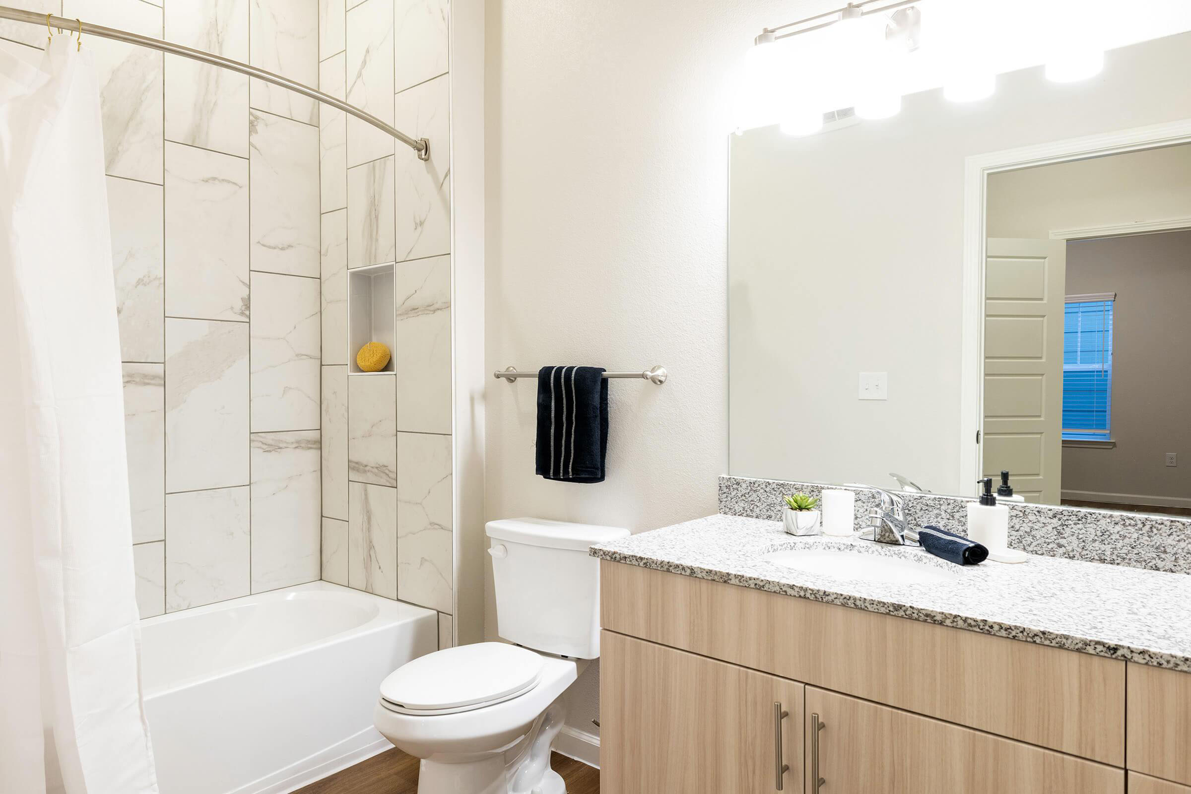 A modern bathroom featuring a white bathtub with a shower curtain, marble-patterned wall tiles, a granite countertop with a sink, and a mirror. There are toiletries on the counter, and a plant decoration. A towel hangs on a silver rack near the sink, and a window lets in natural light.