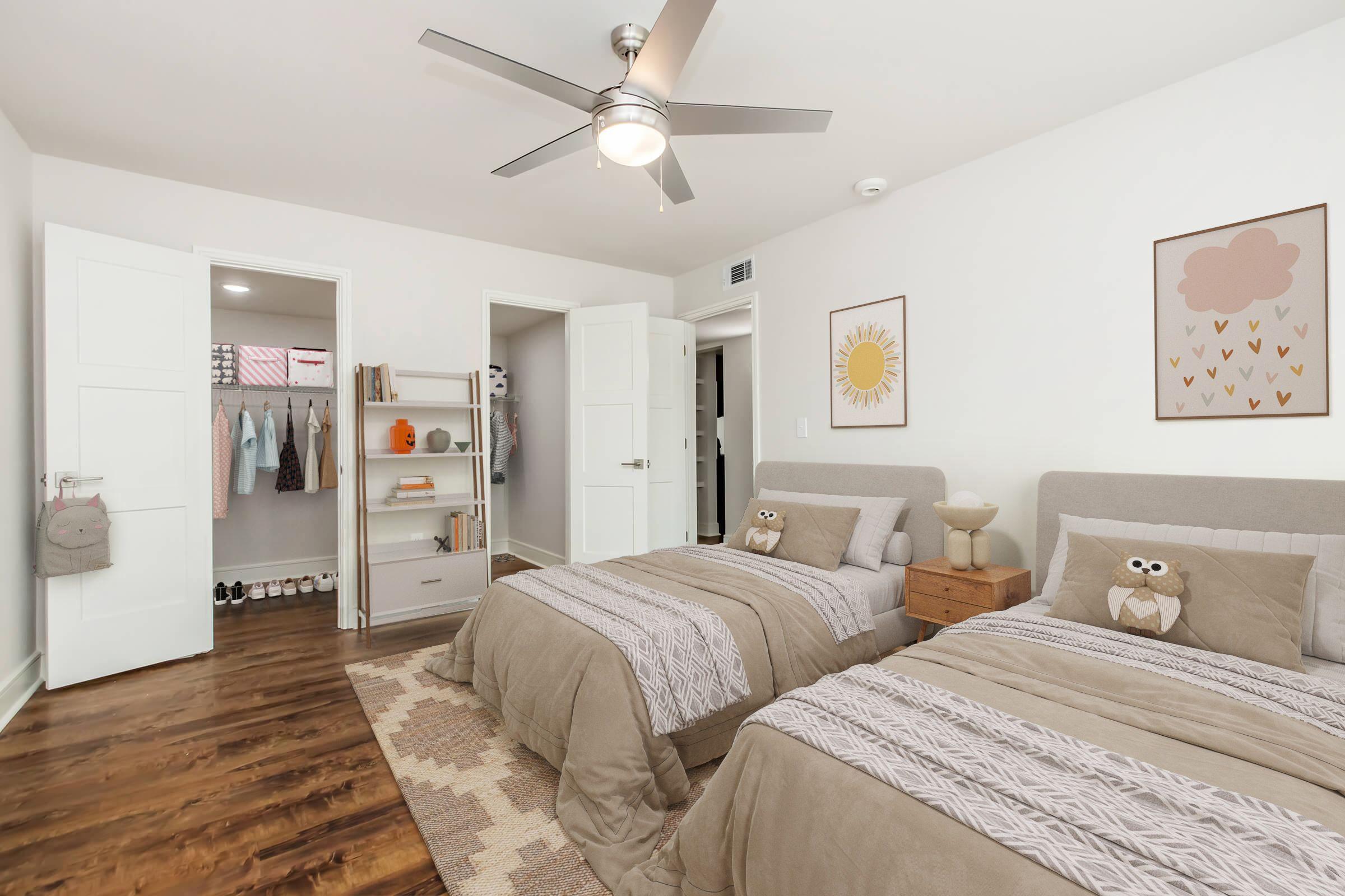 A cozy bedroom featuring two twin beds with gray bedding and owl-themed pillows. The room has light wood flooring, a ceiling fan, and is decorated with framed artwork. A closet with clothes is visible, along with a small shelf holding books and decorative items. A doorway leads to another room.
