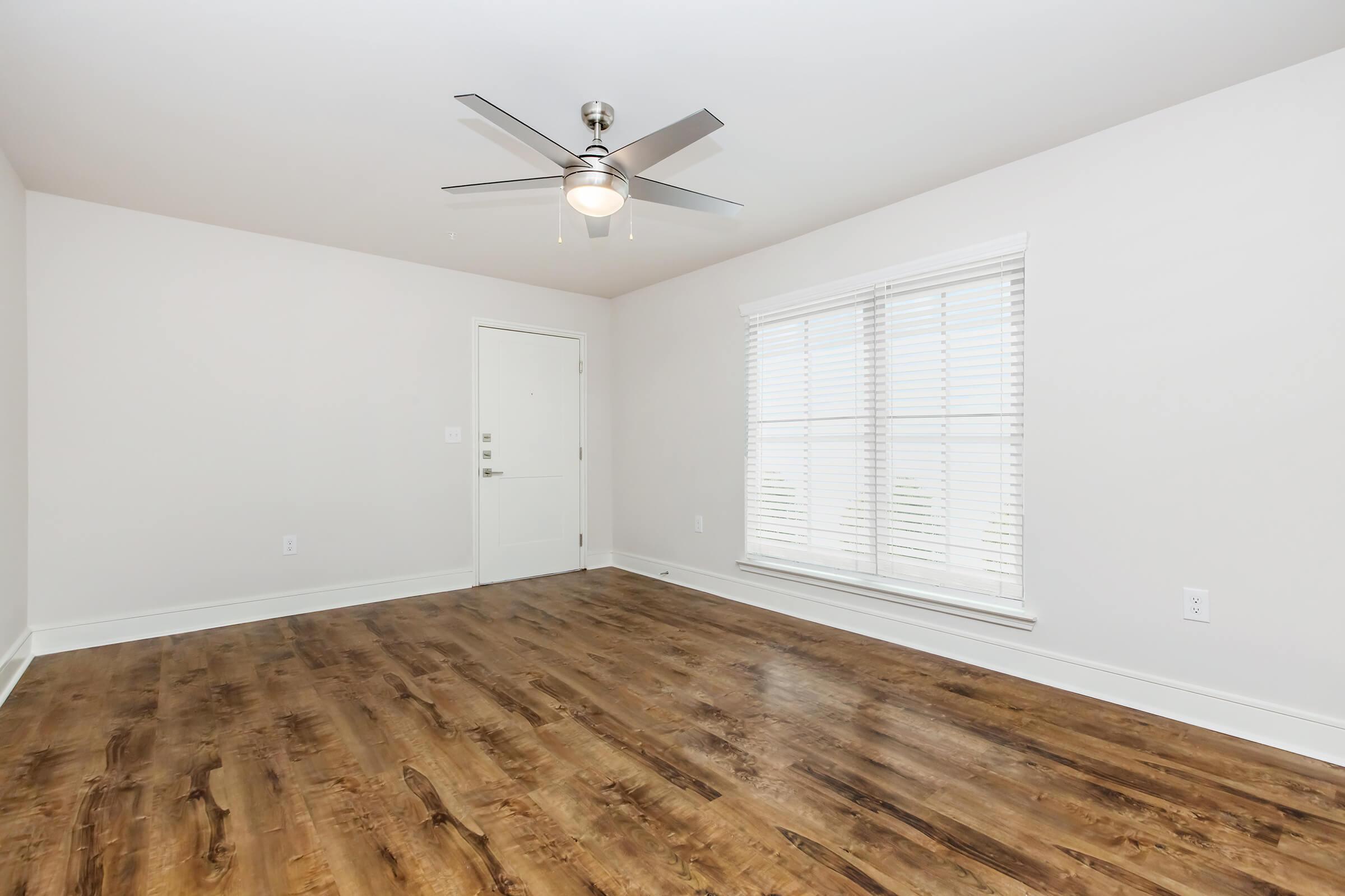A bright, empty room featuring a wooden floor, a ceiling fan with light, and large windows with white blinds. The walls are painted in a light color, and there's a door leading to another area. The overall atmosphere is clean and spacious, ideal for various uses.