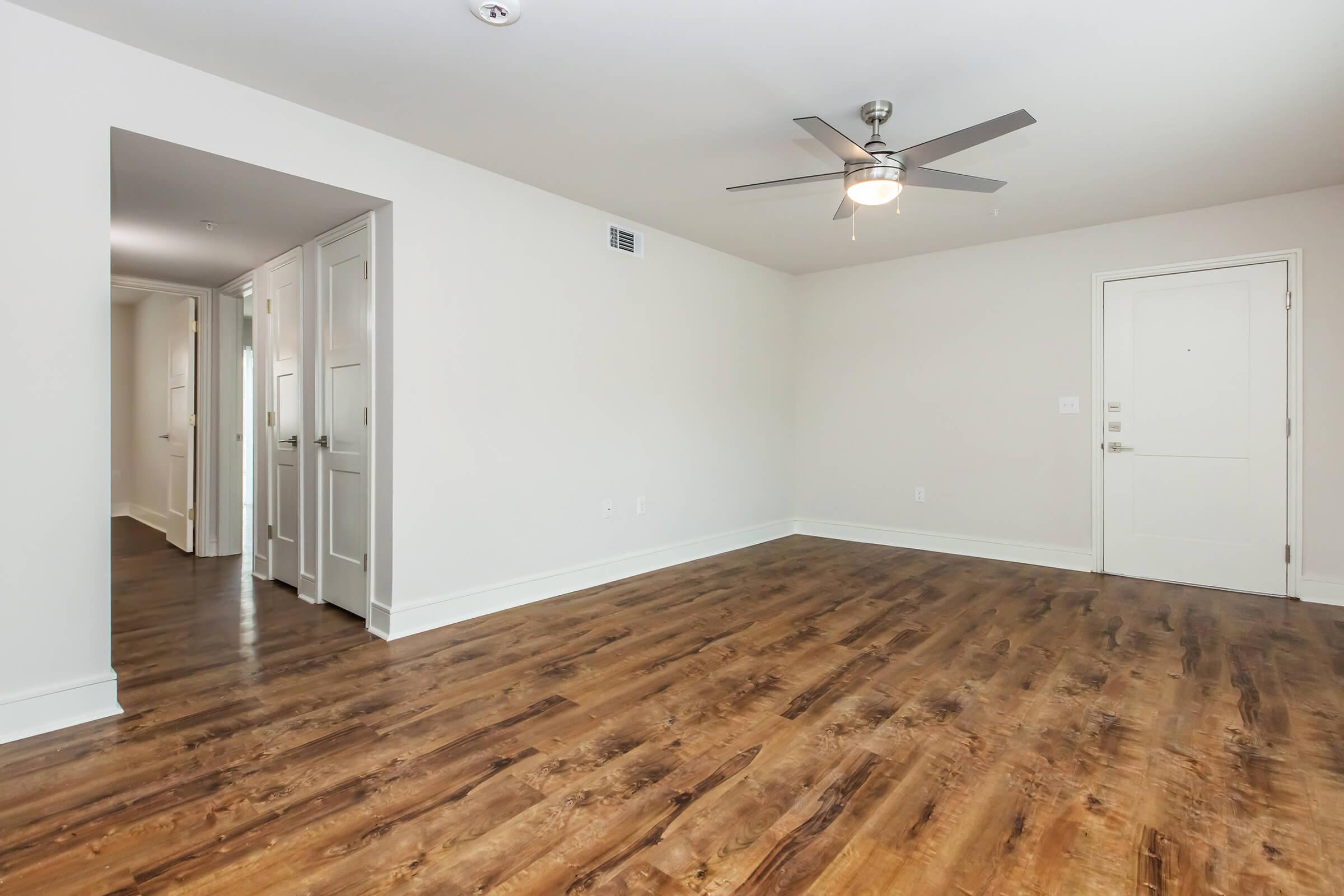 A spacious, empty room with light-colored walls and a polished wood floor. A ceiling fan is installed in the center. To the left, there's an open doorway leading to a hallway. The entrance door is on the right side, with natural light enhancing the welcoming atmosphere.