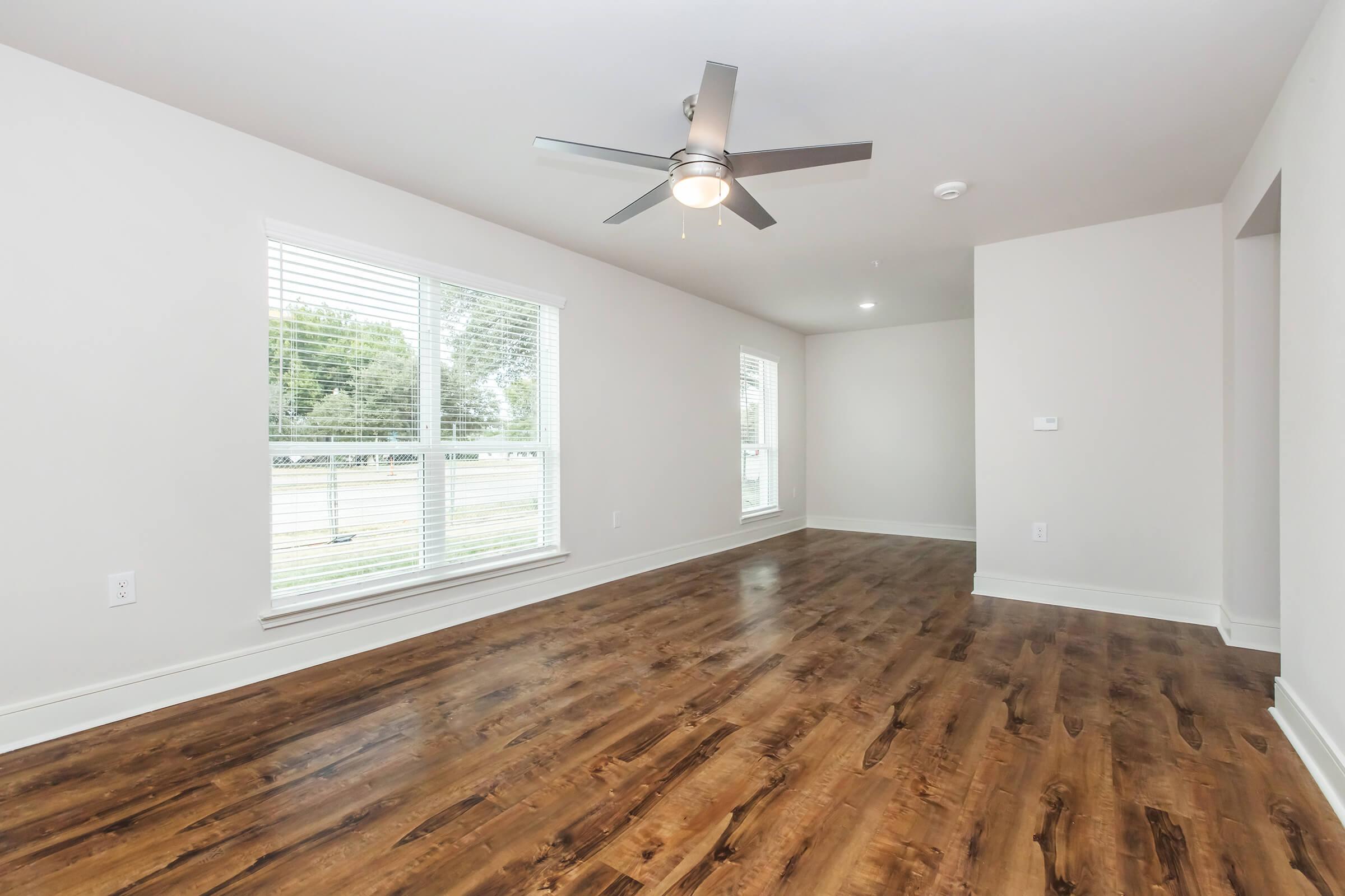 A spacious and bright living room with light-colored walls and a wooden floor. Large windows allow natural light to fill the space, and a ceiling fan is mounted in the center of the room. The overall atmosphere is modern and inviting, providing an open layout.