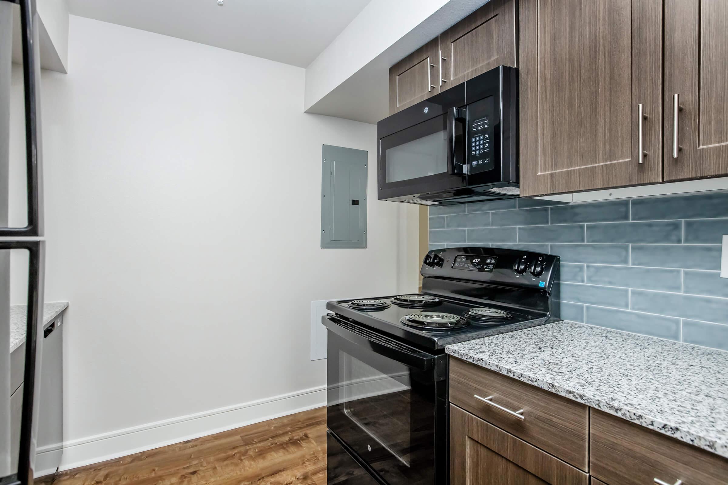 Modern kitchen featuring a black stove and microwave, granite countertops, and wooden cabinetry. The backsplash is made of light blue tiles, and an electrical panel is mounted on the wall. The flooring is light wood, creating a stylish and contemporary atmosphere.