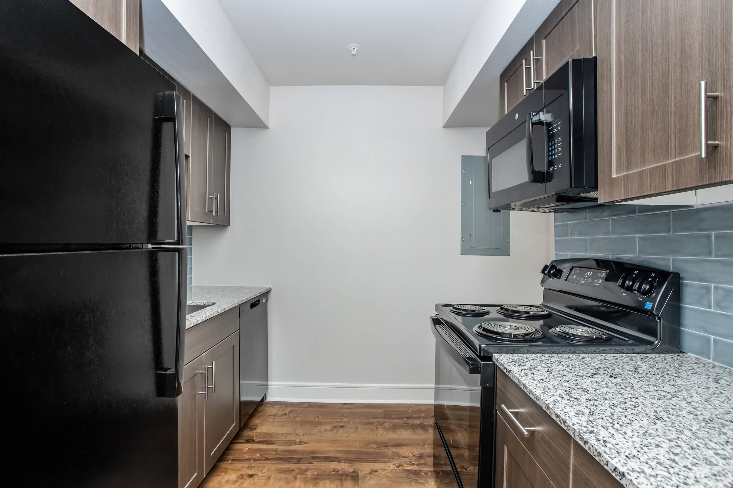 Modern kitchen featuring dark cabinetry, a black refrigerator, and a black microwave above a black stove. The countertops are a light gray with speckles, and the floor is wood. Walls are painted light gray. A window is visible, indicating natural lighting in the space.