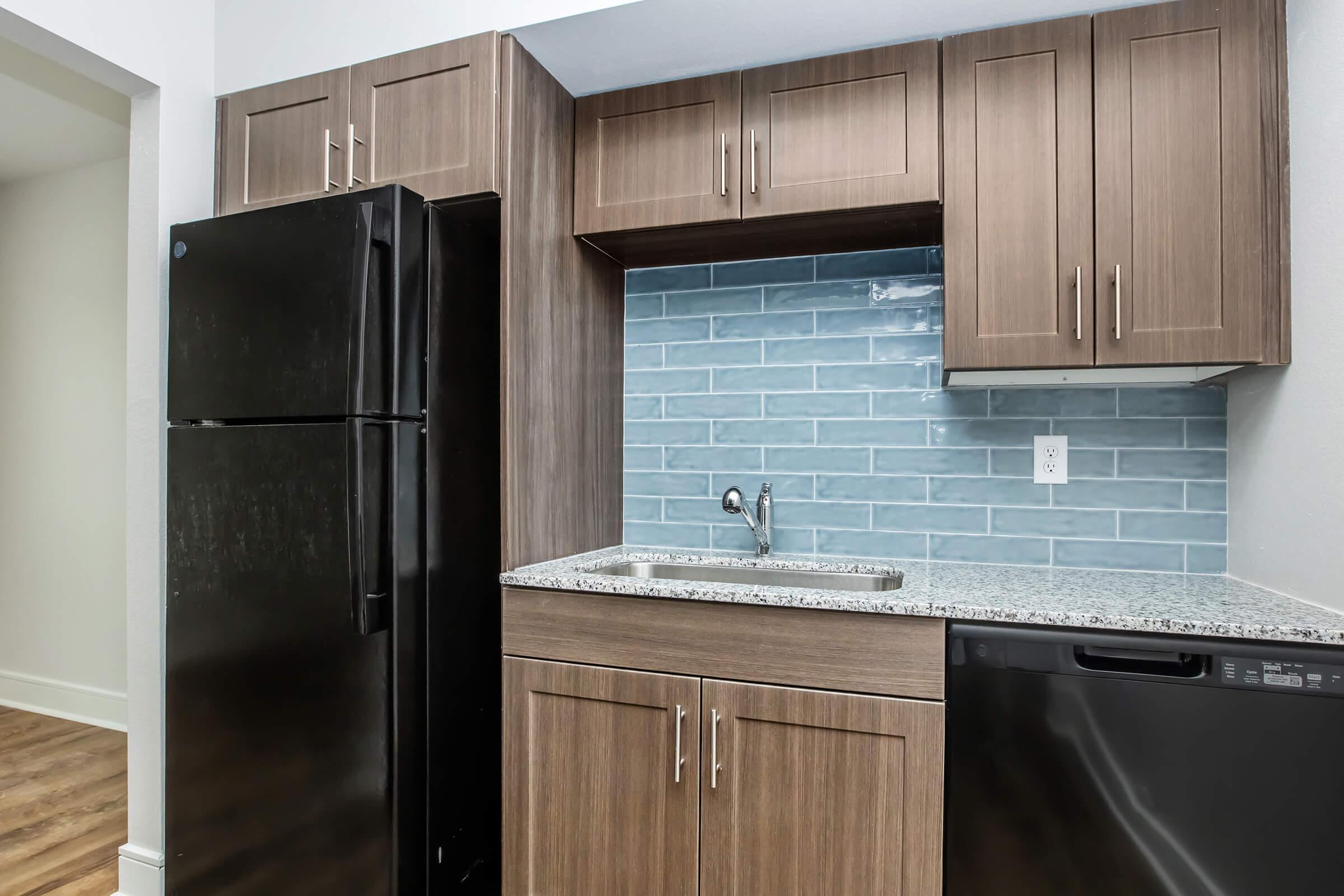A modern kitchen featuring a black refrigerator, dark wood cabinetry, a light blue backsplash, a stainless steel sink, and a granite countertop. The dishwasher is also black, and the flooring is a warm wooden texture. The overall design is sleek and contemporary.