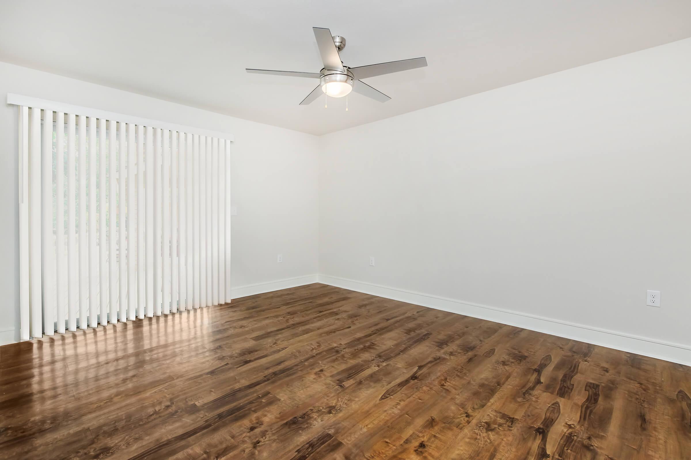 A clean, empty room with light-colored walls and hardwood flooring. A ceiling fan is mounted in the center, and large vertical blinds cover a window on one side, allowing natural light to illuminate the space. The room is spacious and uncluttered, suitable for various uses.