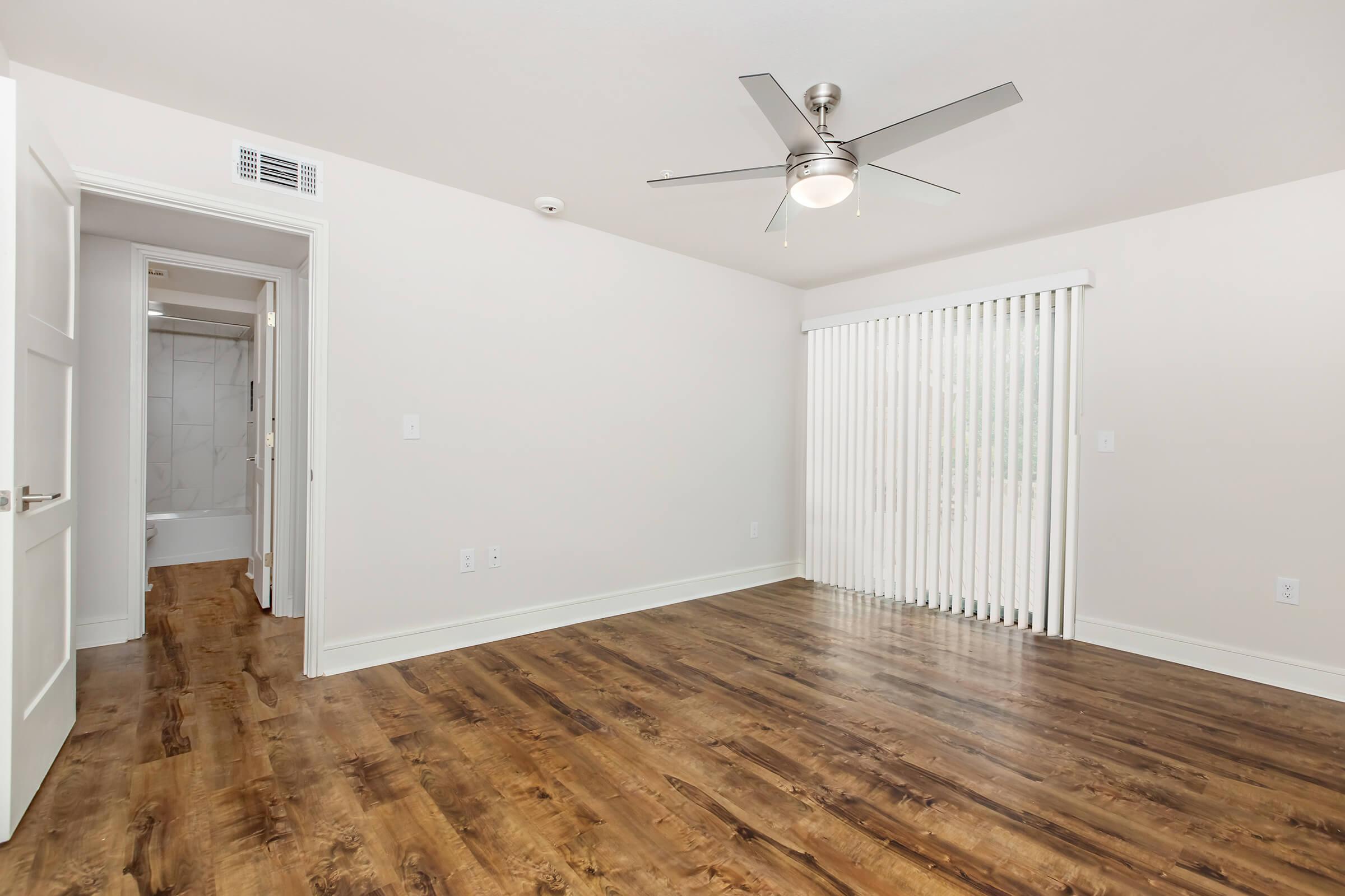 Spacious, modern room with hardwood flooring, a ceiling fan, and large window with vertical blinds. A doorway leads to a bathroom area, featuring neutral-colored walls and a light, airy atmosphere. Ideal for comfortable living or as a cozy study space.