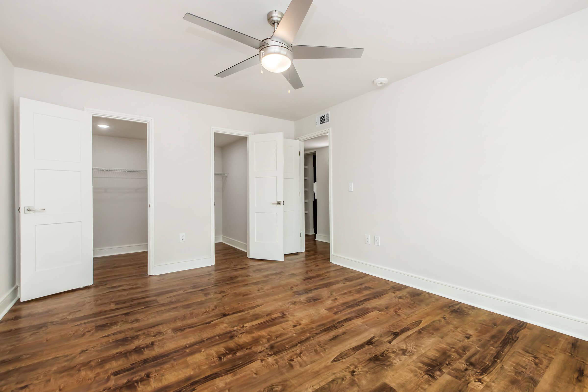 A spacious, well-lit room with light-colored walls and a warm wooden floor. The room features a ceiling fan, two open doors leading to closets, and an overall modern design, creating an inviting atmosphere.