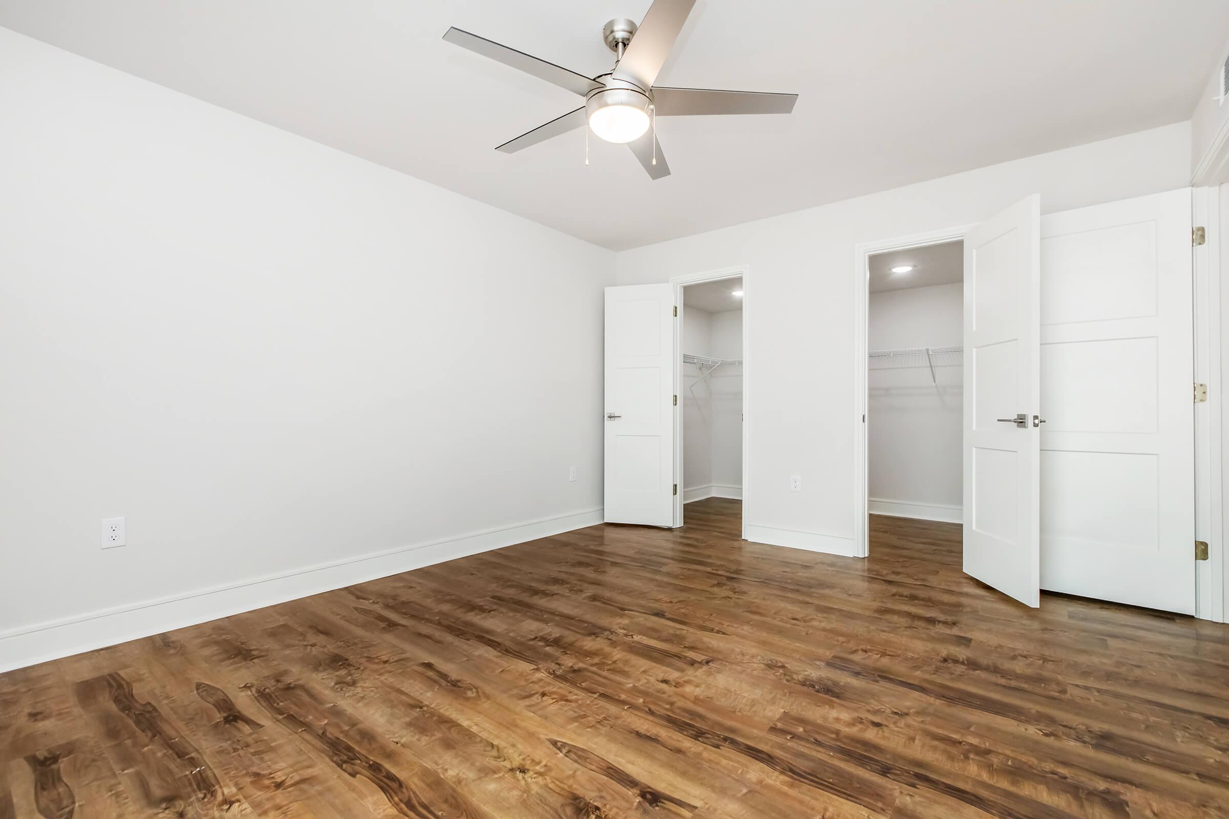 A spacious, empty room with light-colored walls and a polished wooden floor. It features a ceiling fan and two open doors leading to a closet area. The room has a bright, airy feel with ample natural light.