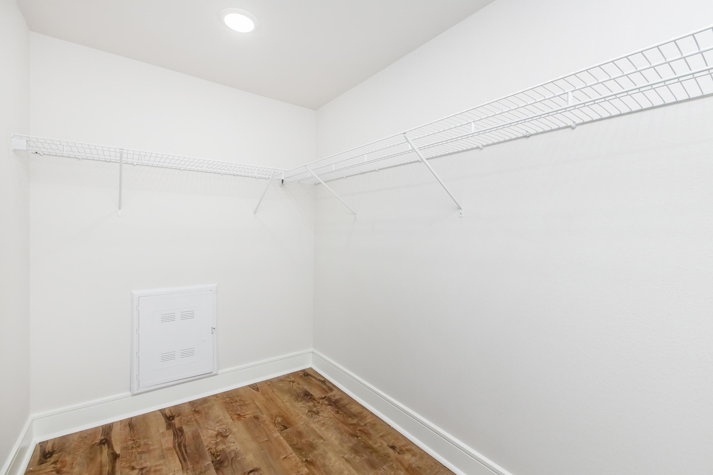 A spacious, empty closet featuring white wire shelving on two sides, with a small access panel on the wall. The floor is finished in wood, and the walls are painted white. A ceiling light illuminates the space, creating a clean and organized appearance.