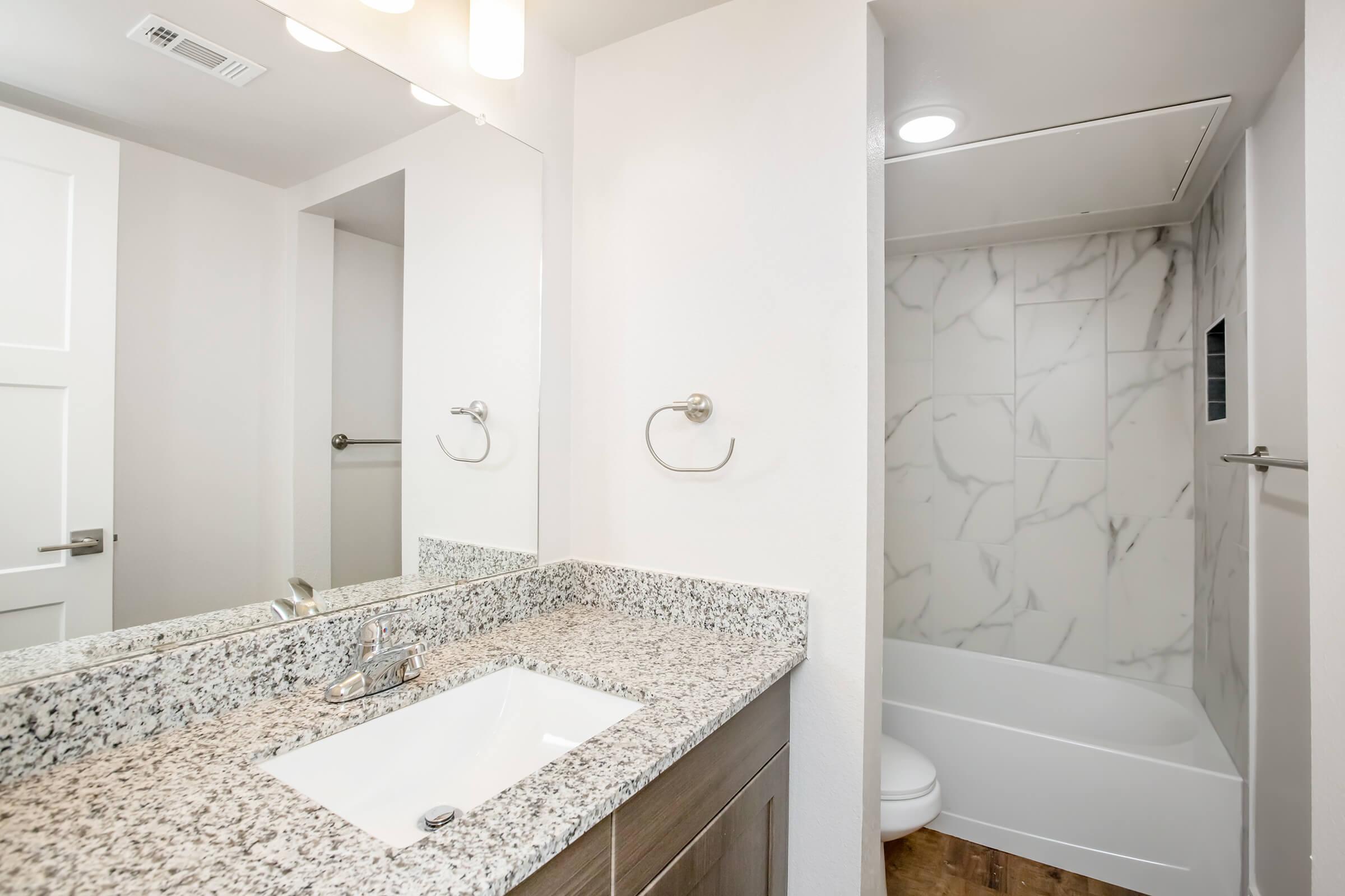 A modern bathroom featuring a white sink with a granite countertop, a mirror above the sink, and a shower/bath area with marble-patterned tiles. The walls are painted in a light color, and there is a towel bar beside the mirror. Overall, the space is clean and minimalist in design.