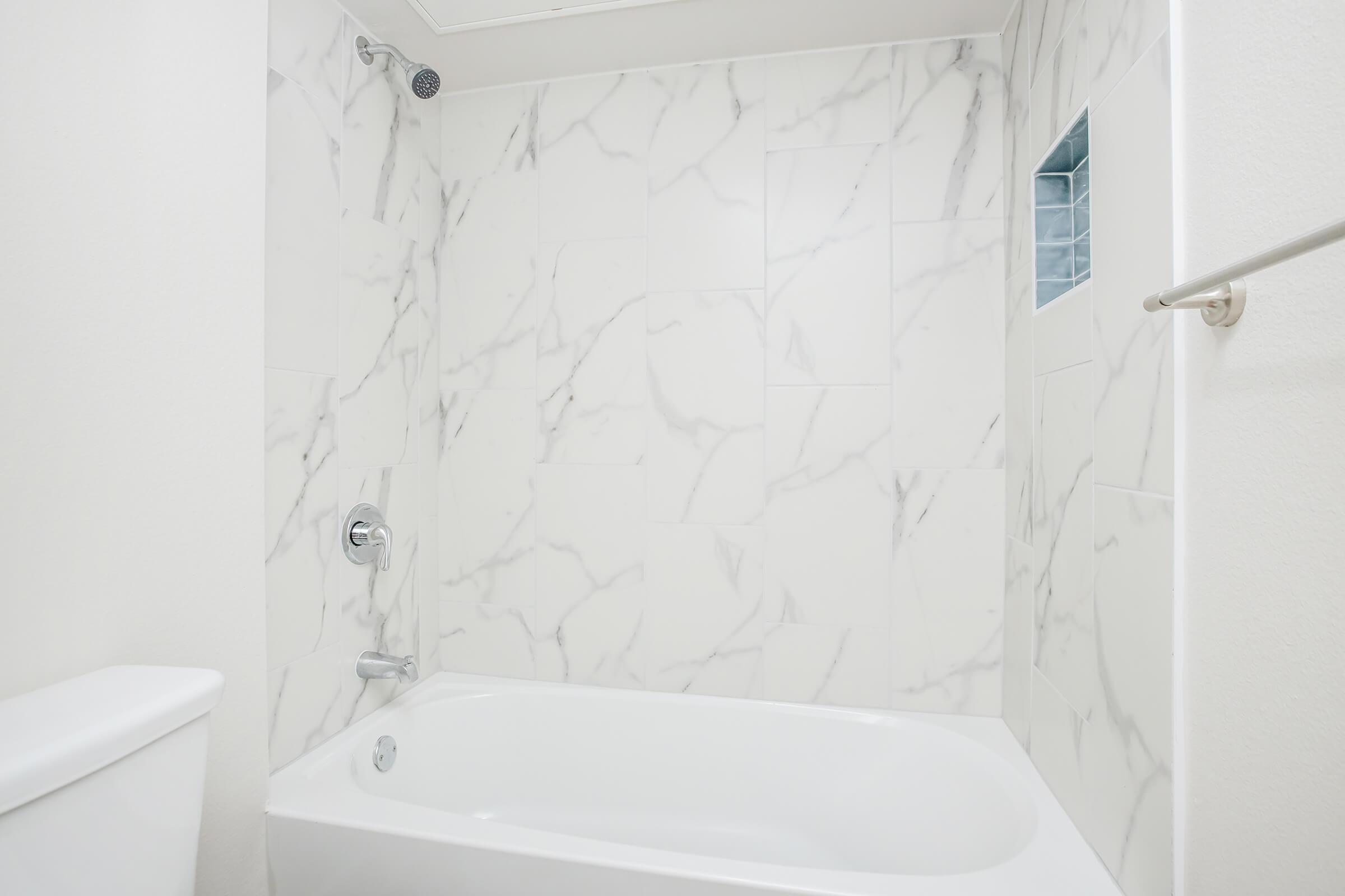 A clean bathroom featuring a white bathtub with a marble-patterned wall. The showerhead is mounted above, and a small window is set into the wall, allowing natural light to enter the space. The overall design is modern and minimalist.