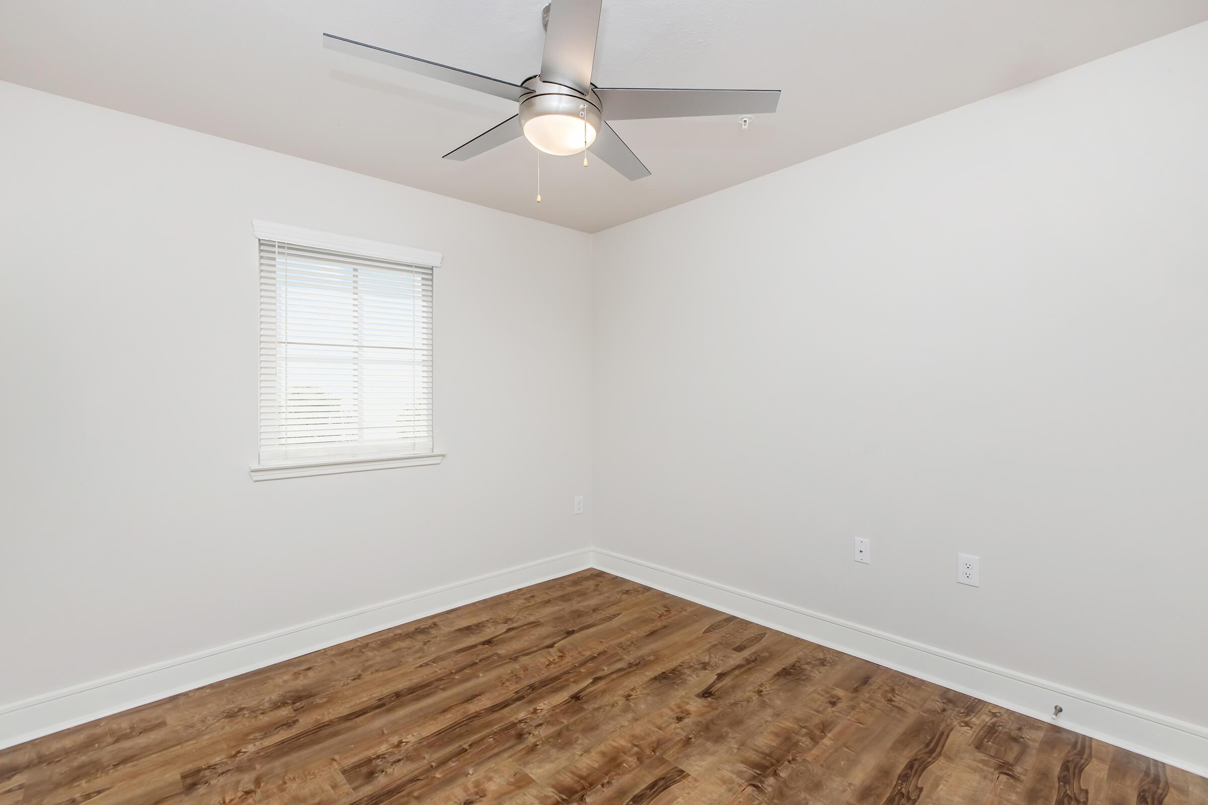 Empty room with light-colored walls and a wooden floor. A ceiling fan with a light fixture is mounted in the center, and there is a window with white blinds on one wall, allowing natural light to enter. The overall appearance is clean and modern, ready for furnishing.