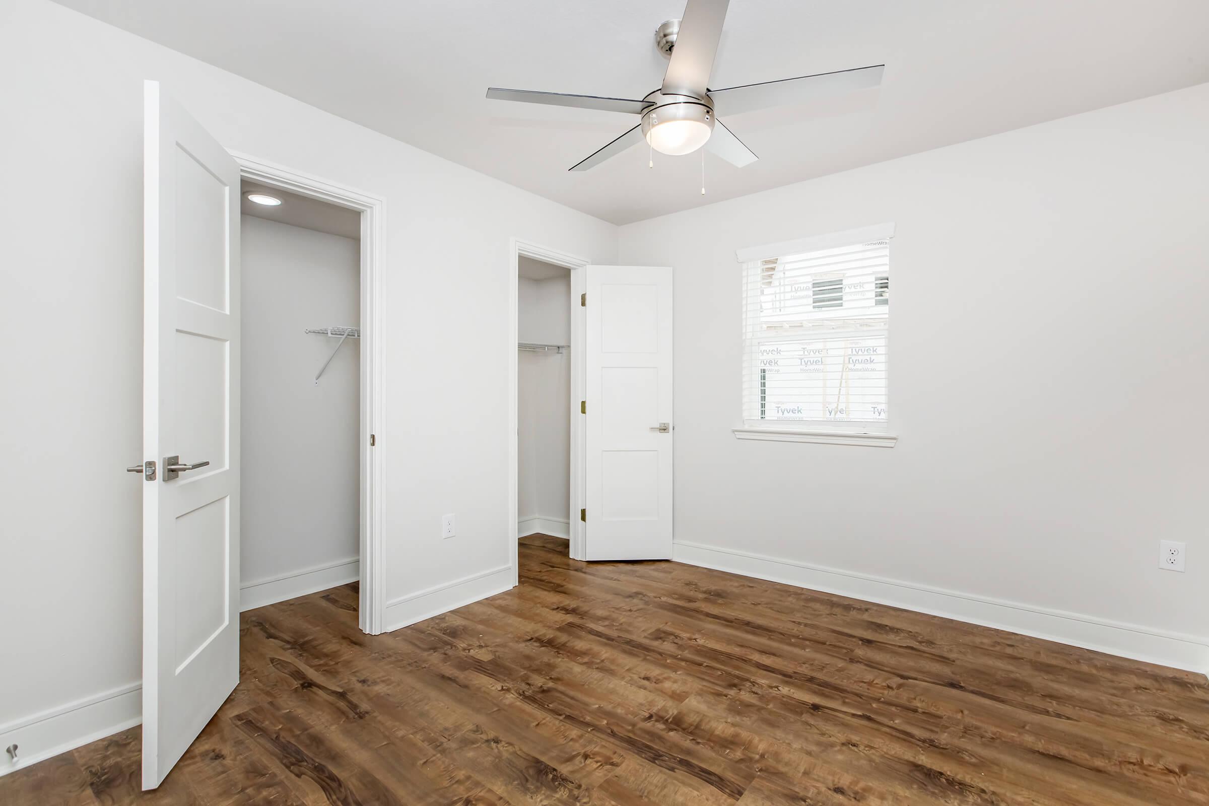 A well-lit, empty bedroom featuring light gray walls, a ceiling fan, and a window with blinds. There are two doors: one leading to a closet and another to an adjoining room, with polished wooden flooring throughout. The room has a clean and modern appearance.