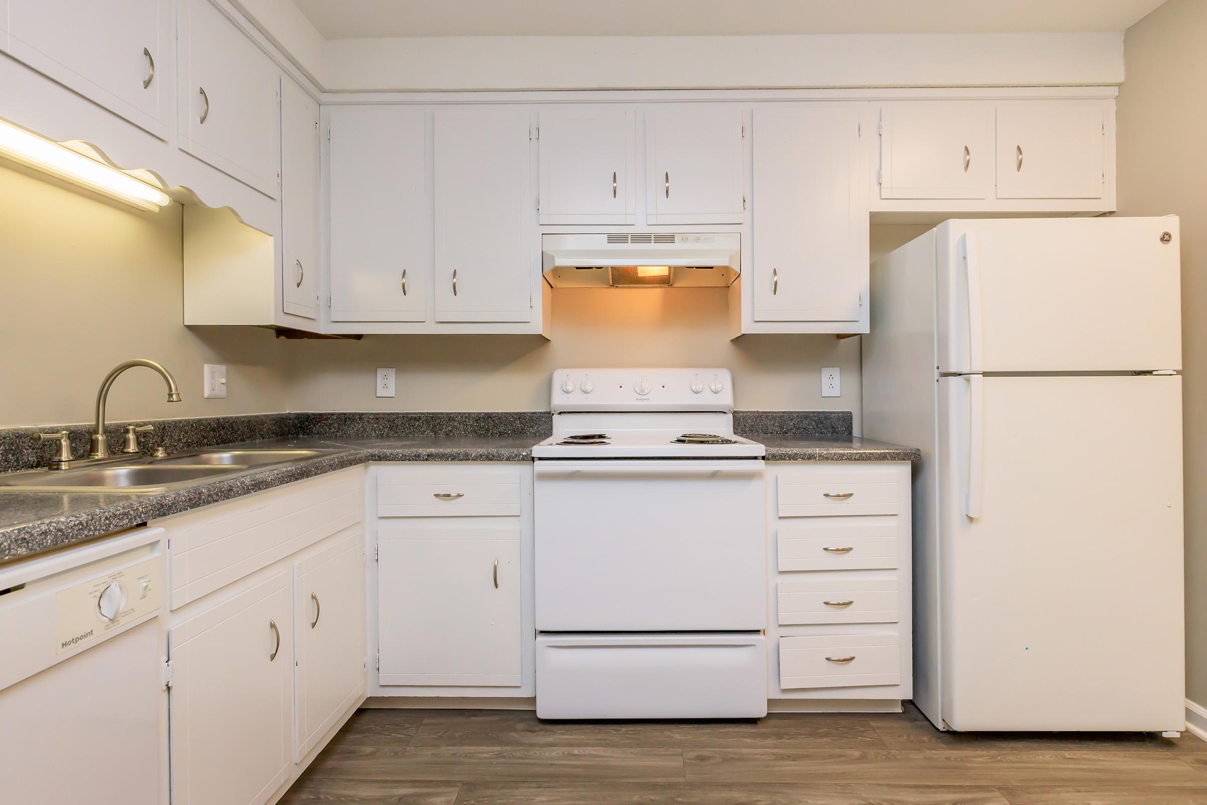 A clean, modern kitchen featuring white cabinetry, a stainless steel sink, a white stove and oven with a range hood, and a white refrigerator. The countertops are dark granite, and the walls are painted a soft light color, creating a bright and inviting space.