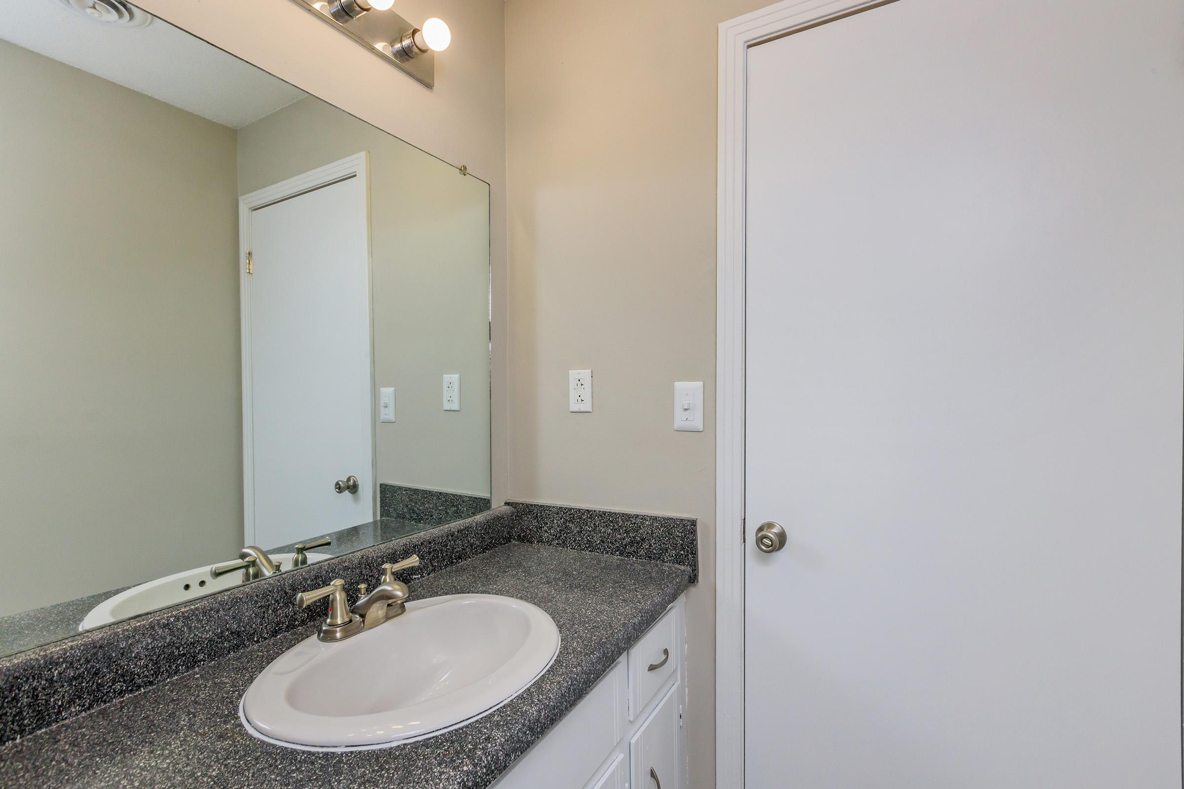 A bathroom interior featuring a mirrored wall above a granite countertop with a sink and faucet. There are light fixtures above the mirror, and a white door is visible in the background. The walls are painted a neutral color, creating a clean and modern look.