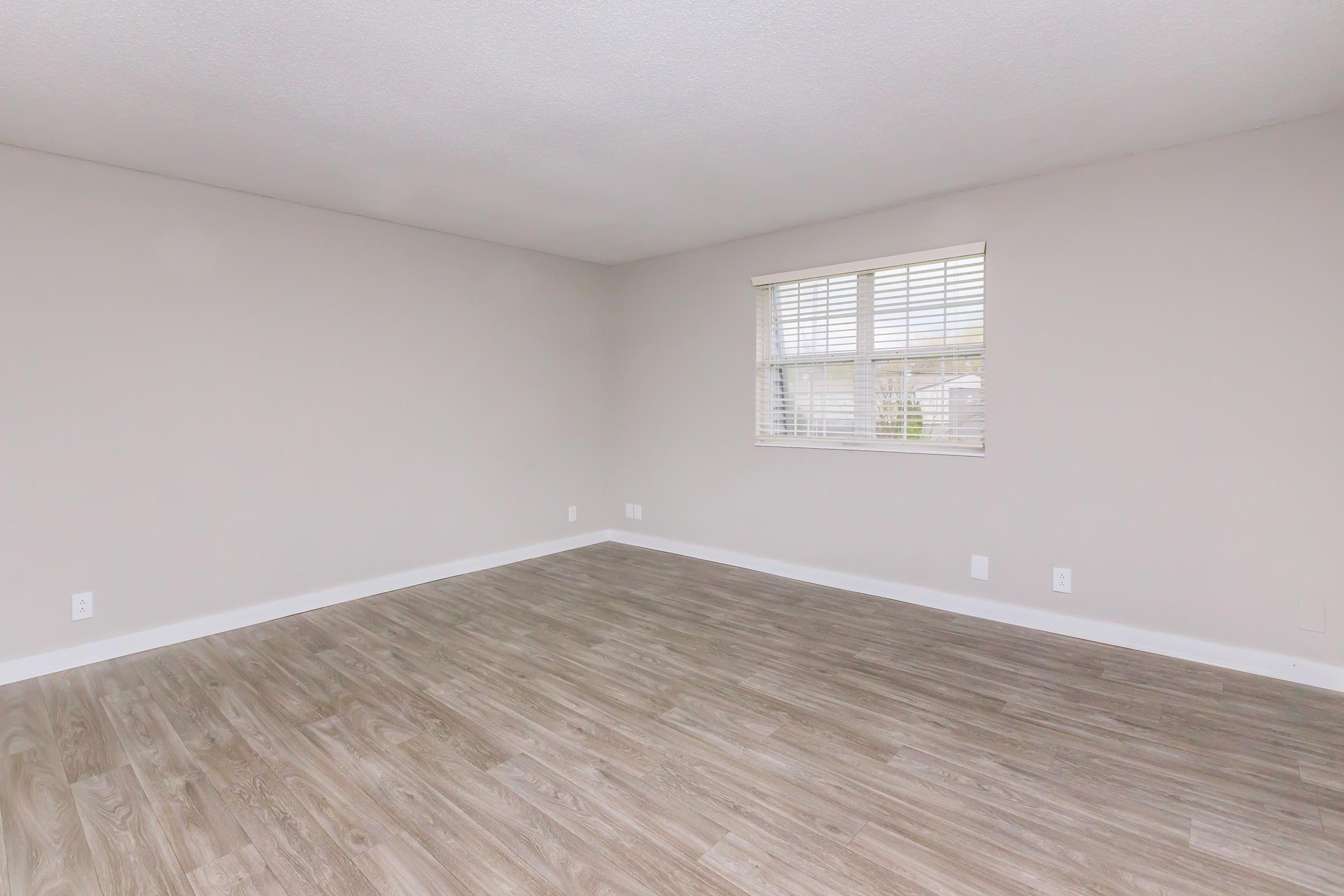 Empty room with light-colored walls and a window with blinds. The flooring is a wooden laminate, and the space is well-lit but devoid of furniture or decorations, creating a minimalist atmosphere.