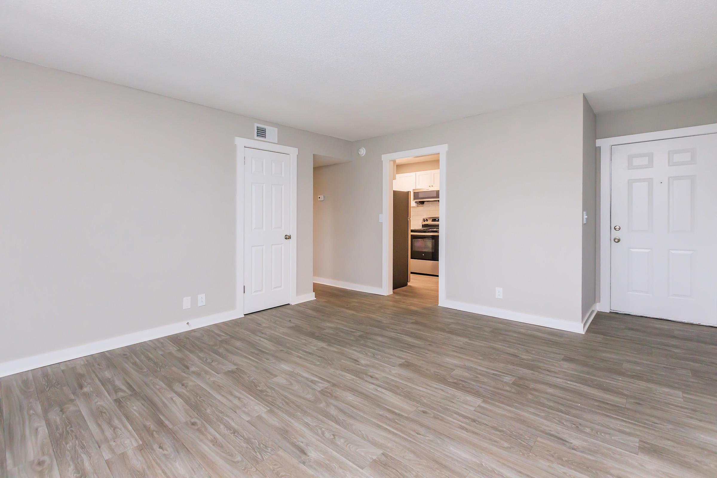 A spacious, unfurnished room with light gray walls and a wooden floor. Two closed doors on the sides lead to other areas, and a view of a kitchen can be seen through an opening. Natural light illuminates the room, creating a bright and airy atmosphere.