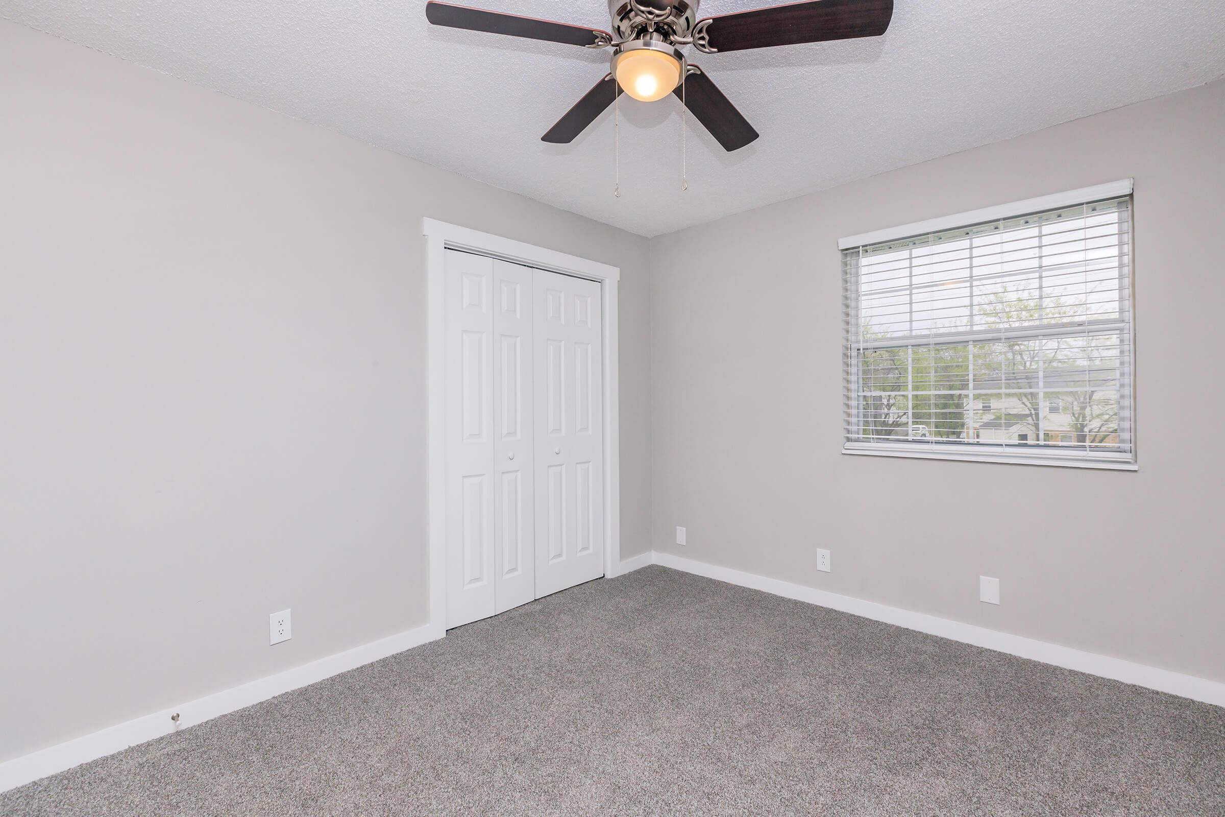 A well-lit, empty bedroom featuring light gray walls and a ceiling fan. The floor is covered with a soft, light-colored carpet. There's a window with blinds allowing natural light in, and a white double door closet situated on the wall opposite the window.