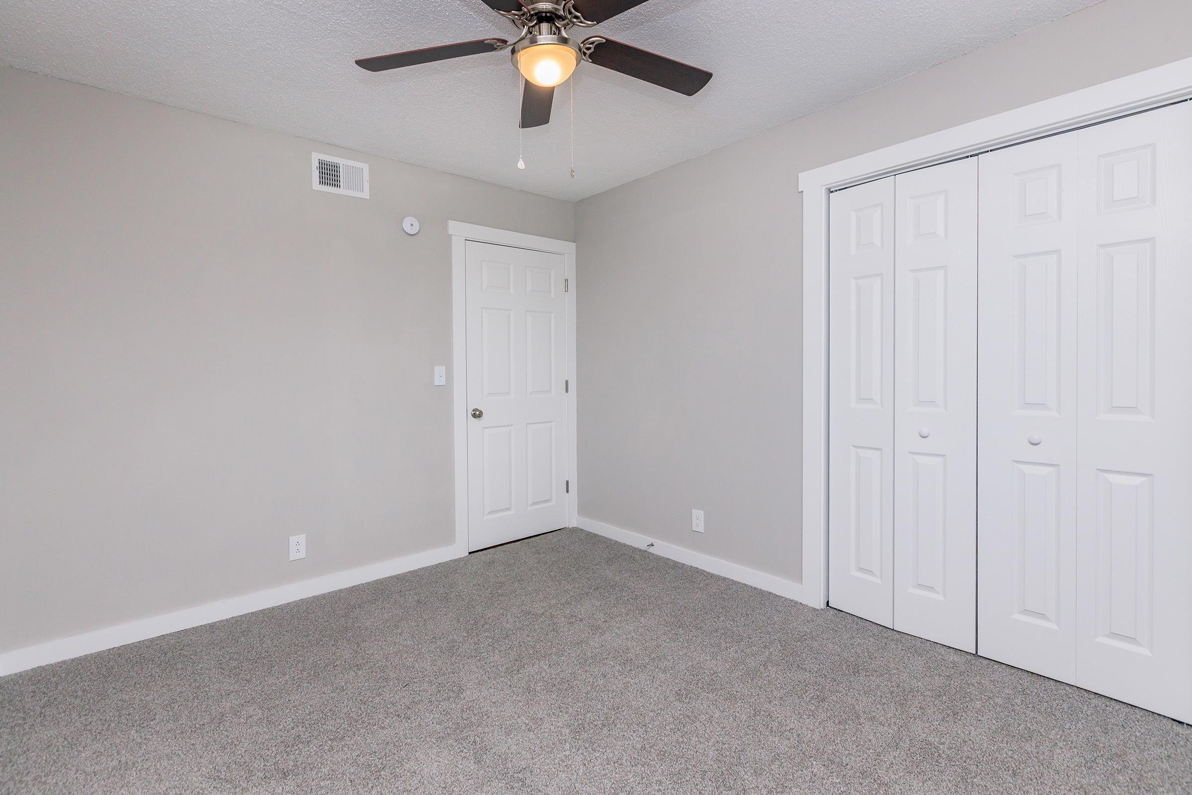 A spacious room with light gray walls and carpet. A ceiling fan is installed, and there is a white door on one wall along with double closet doors featuring white panels. The room is well-lit, offering a clean and airy atmosphere.
