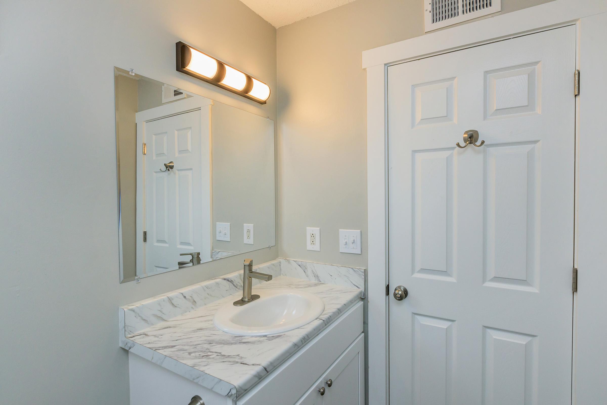 A modern bathroom featuring a white vanity with a marble countertop and a sink, a large mirror above the sink, and a light fixture mounted on the wall. There is a closed white door and neutral-colored walls, creating a clean and bright atmosphere.