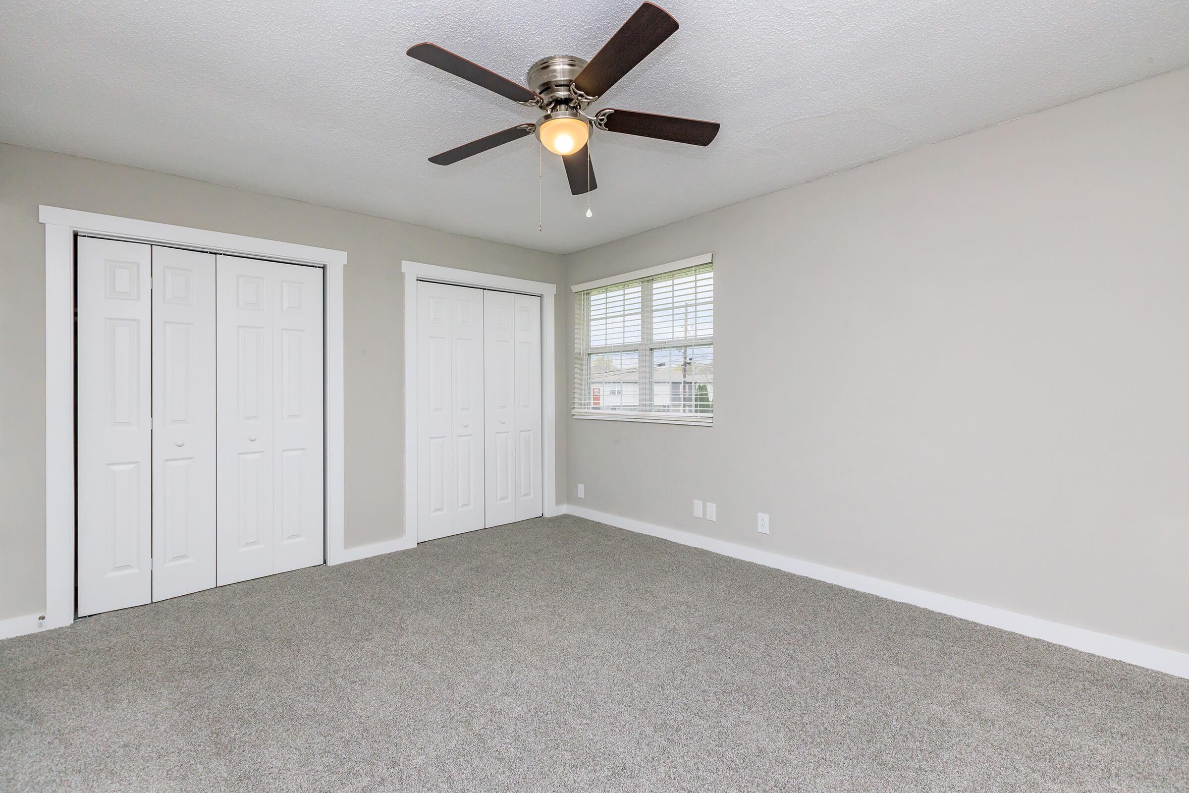 A light and spacious room featuring a ceiling fan, beige walls, and carpeted flooring. Two white double closets are visible on one side, and a window with blinds allows natural light to enter, creating a bright atmosphere in the space.