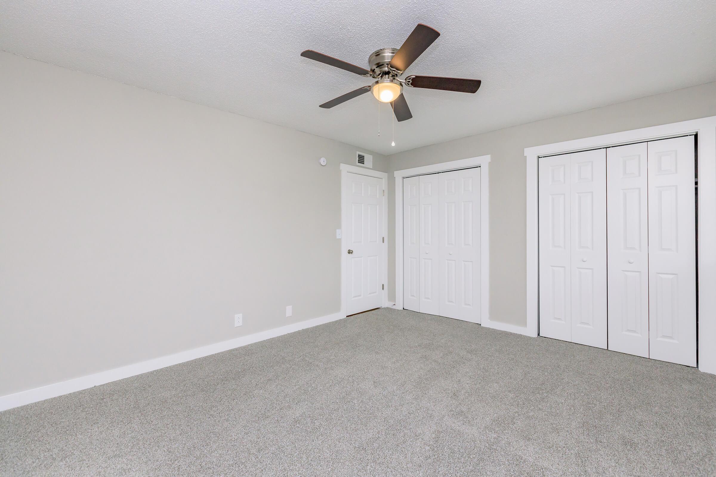 A spacious, light-colored room with a textured ceiling and a ceiling fan. The floor is covered with soft grey carpet. There are two white double closet doors on the right and a doorway to the left. The walls are painted in a neutral tone, creating a clean and inviting atmosphere.