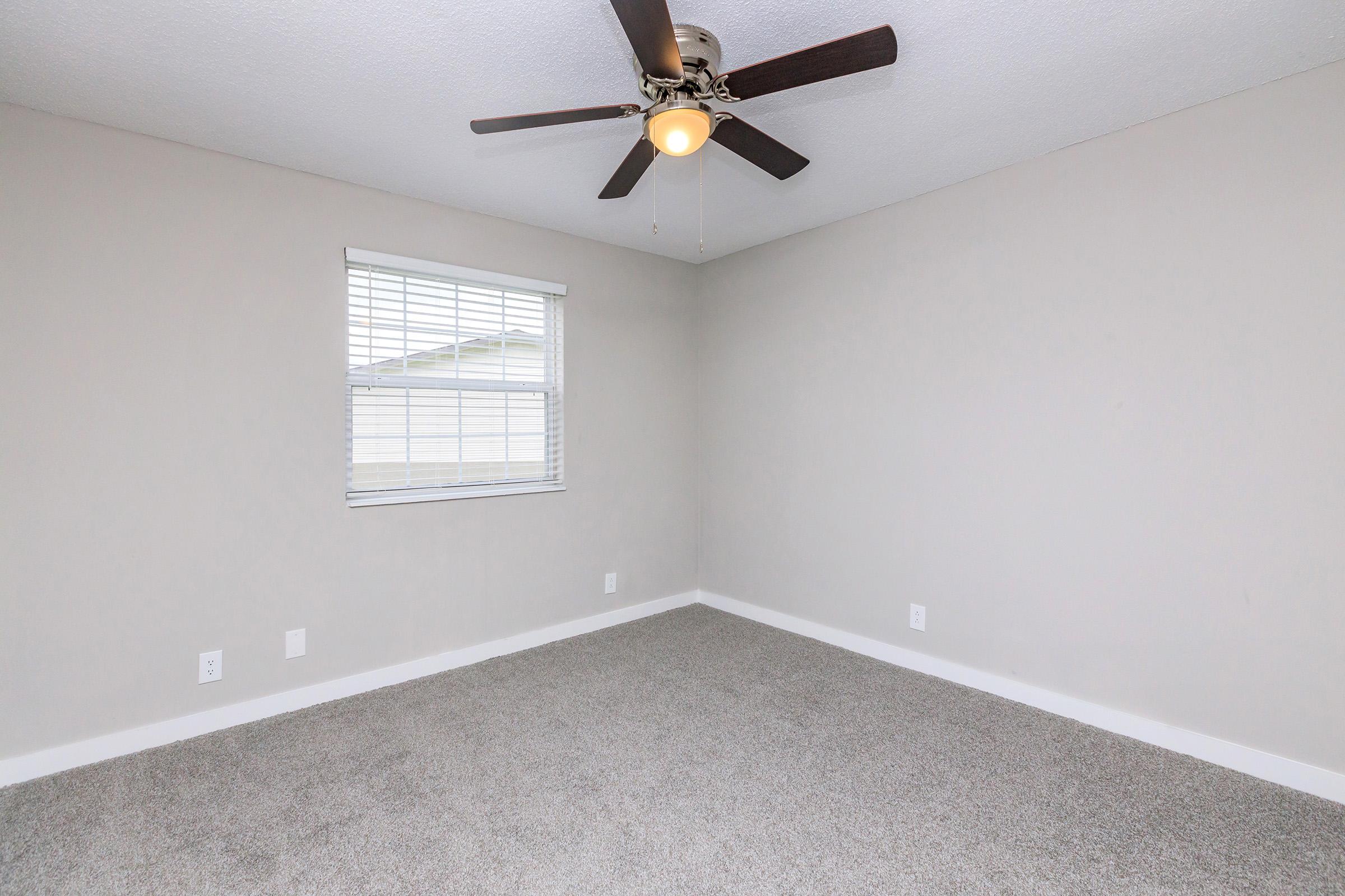 A well-lit empty room with beige walls and a light-colored carpet. It features a ceiling fan with wooden blades and a single window with white blinds allowing natural light to enter. The space is simple and uncluttered, ideal for personal decoration or furnishing.