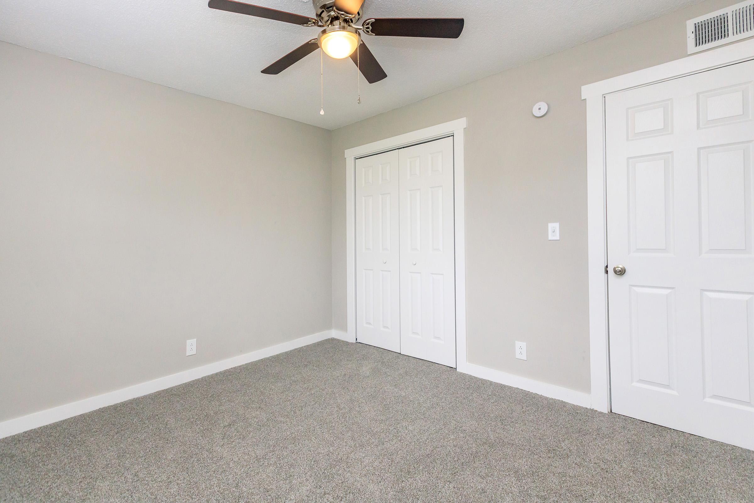 A simple, empty room featuring light gray walls and a ceiling fan. The floor is covered in plush gray carpet. On one side, there are double white doors leading to a closet, and a single white door on the other side. Natural light may enter through a window not visible in the image.