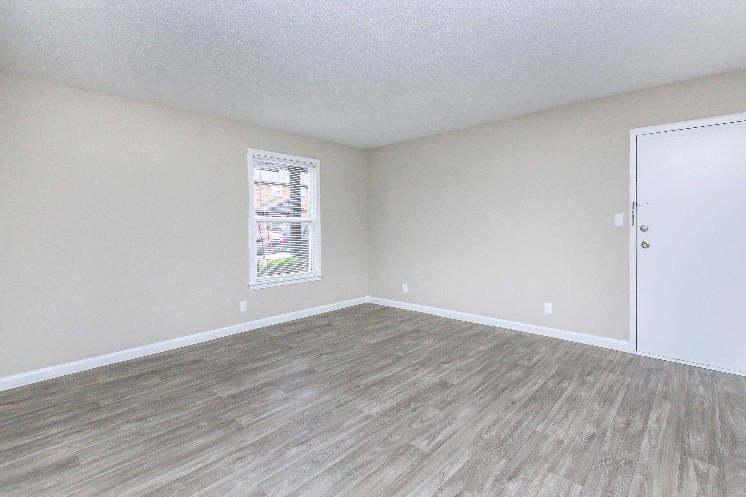 Empty room with light-colored walls and hardwood-style flooring. There is a window on the left side, allowing natural light, and a white door to the right. The space appears clean and well-maintained, suitable for various interior design possibilities.