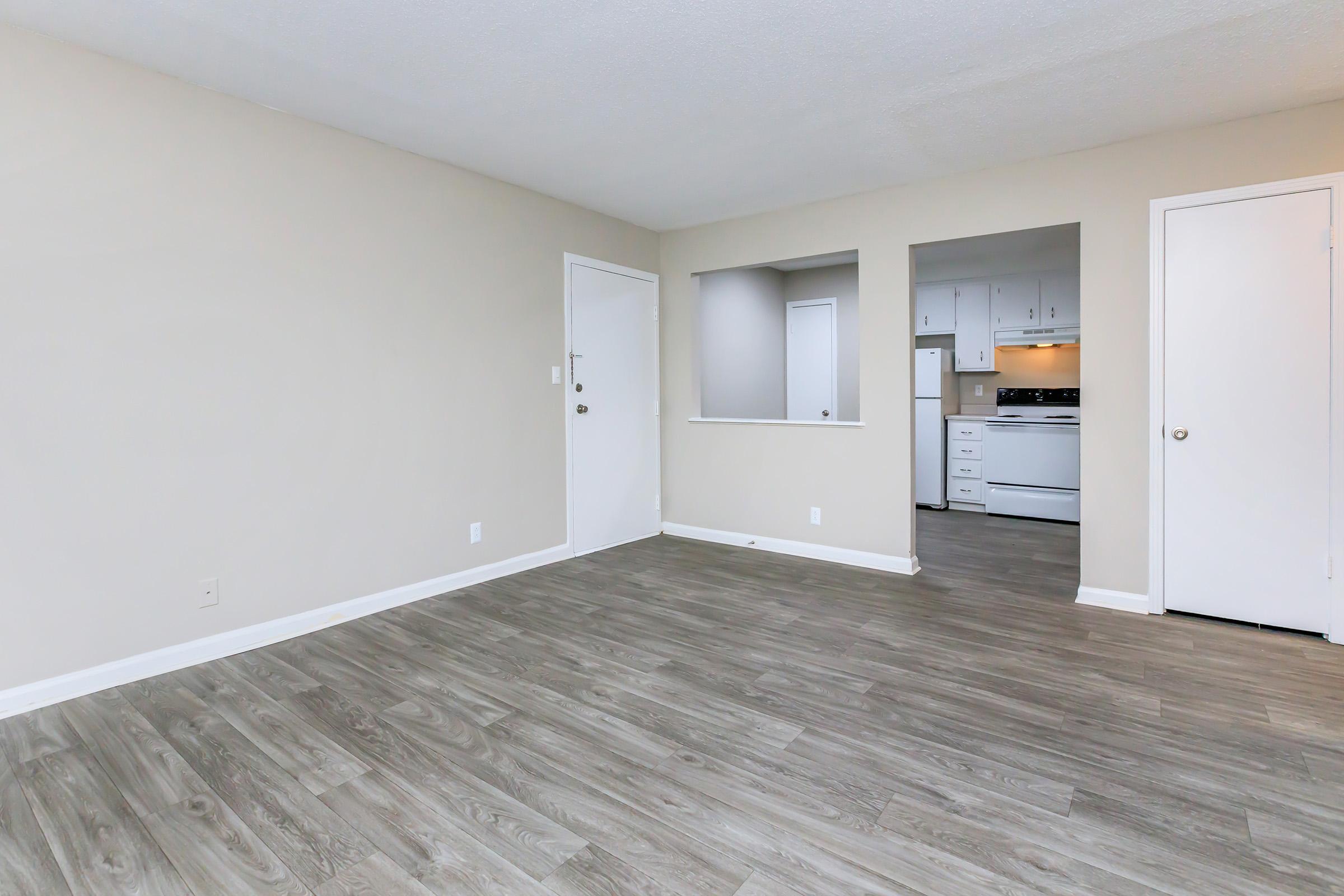 Interior view of a simple, unfurnished living space featuring light-colored walls and a wood-look floor. A doorway leads to a kitchen area with white cabinets and appliances visible in the background. The room is brightly lit and has a neutral color palette, creating an open and airy atmosphere.