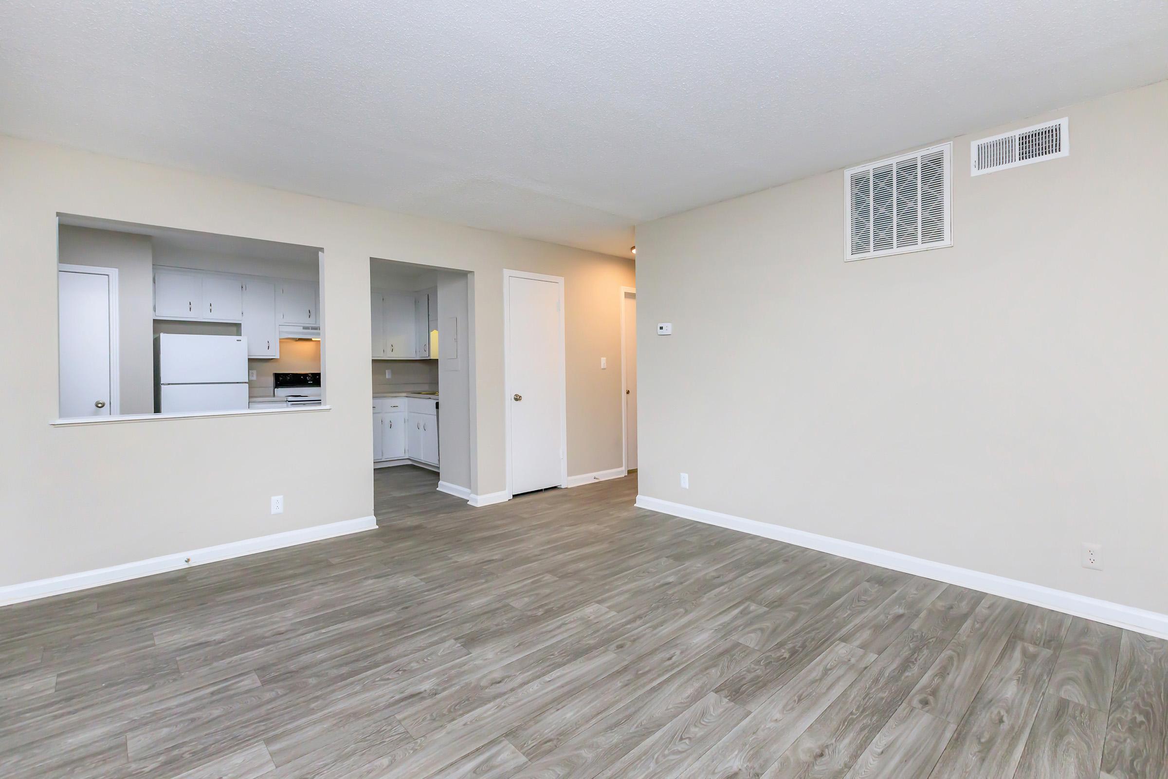 A spacious, empty living room with light-colored walls and a wood-like floor. There is a small open kitchen area visible on the left with white cabinetry. A doorway leads to another room, and a wall vent can be seen on the right. The overall feel is bright and minimalistic.