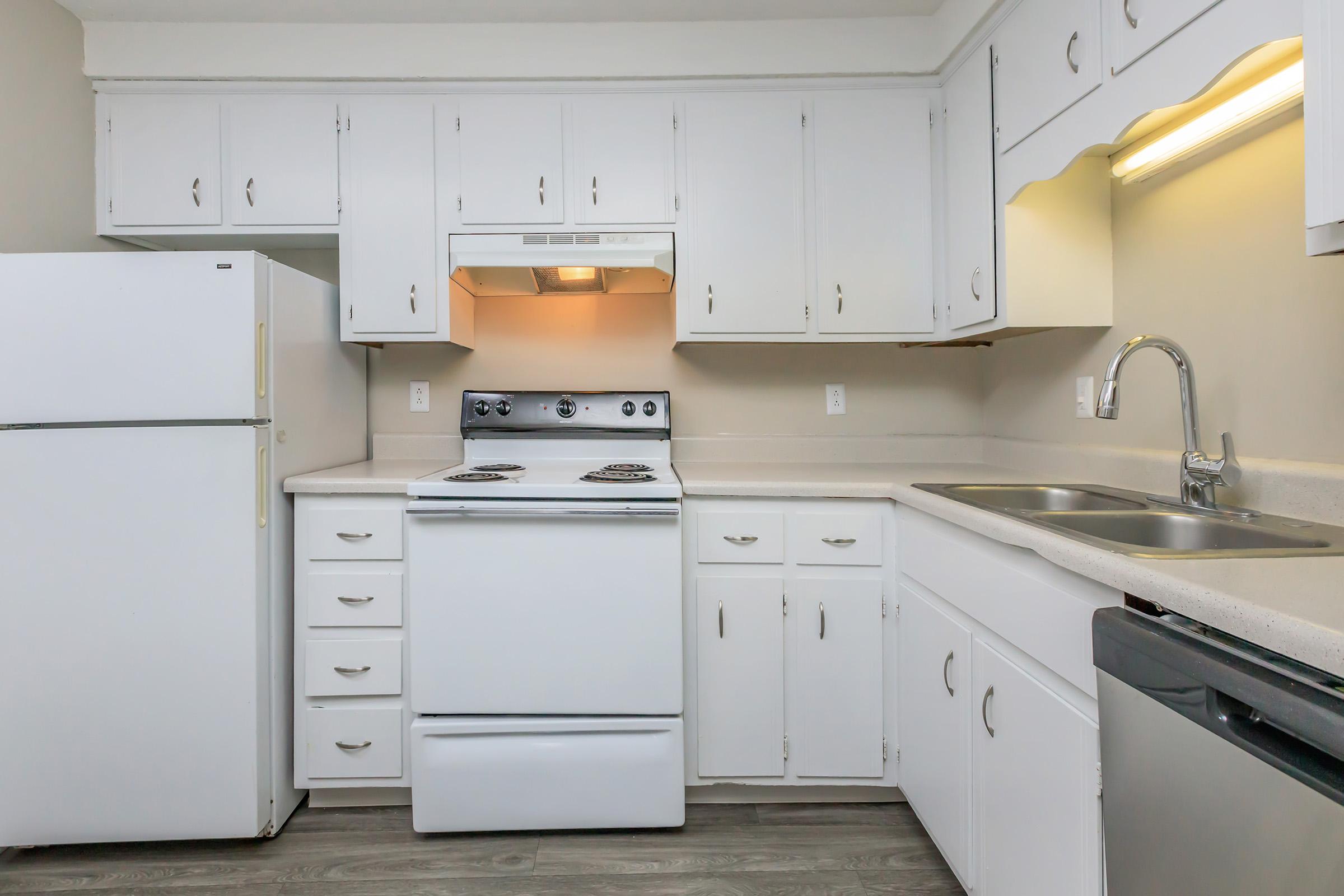 A modern kitchen featuring white cabinets, a white refrigerator, a white stove with an oven, a double sink, and stainless steel appliances. The counter has a light-colored surface and the walls are painted in a neutral tone, creating a clean and bright atmosphere.