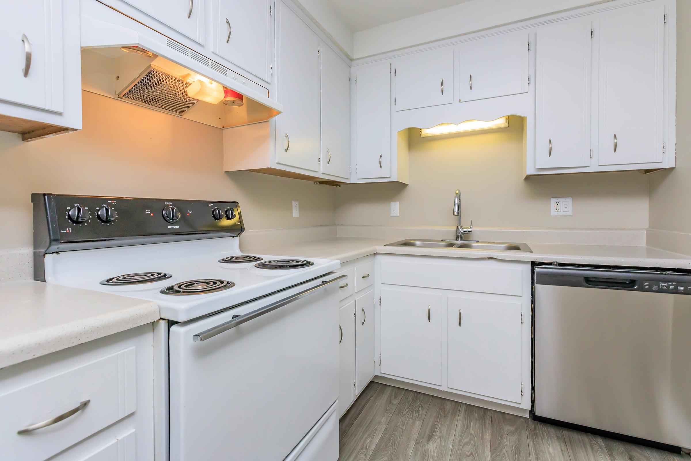 Bright, modern kitchen featuring white cabinetry, a silver stove with four burners, a stainless steel dishwasher, and a double sink. The countertops are light-colored, and the flooring is a wood-like laminate. Adequate lighting is provided by an overhead fixture and under-cabinet lights.
