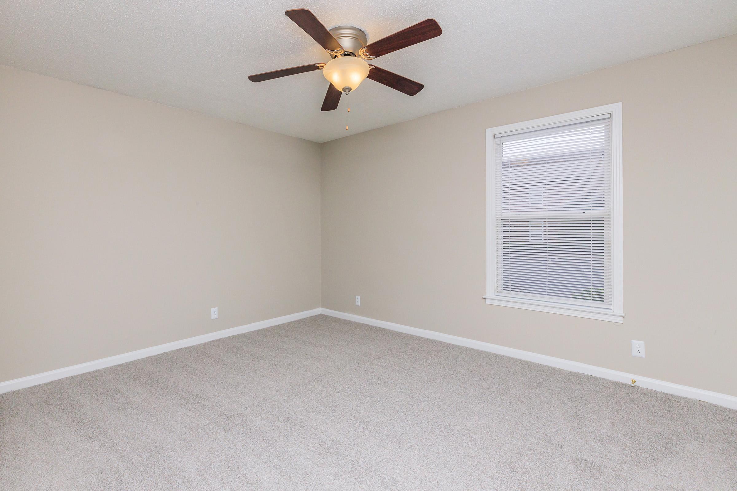 A spacious, empty room with light beige walls and plush carpet. A ceiling fan with wooden blades is centered overhead, while a window with blinds allows natural light to enter. The room is free of furniture, creating an open and airy atmosphere.
