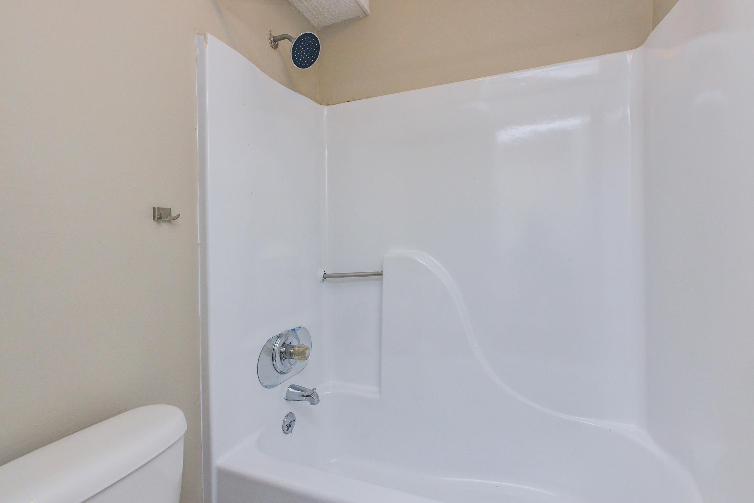 A clean, white bathtub with a showerhead mounted on the ceiling, set against light-colored walls. A chrome faucet and handle are visible on the tub, and a toilet is partially seen on the left side of the image. The overall ambiance conveys a simple, modern bathroom design.