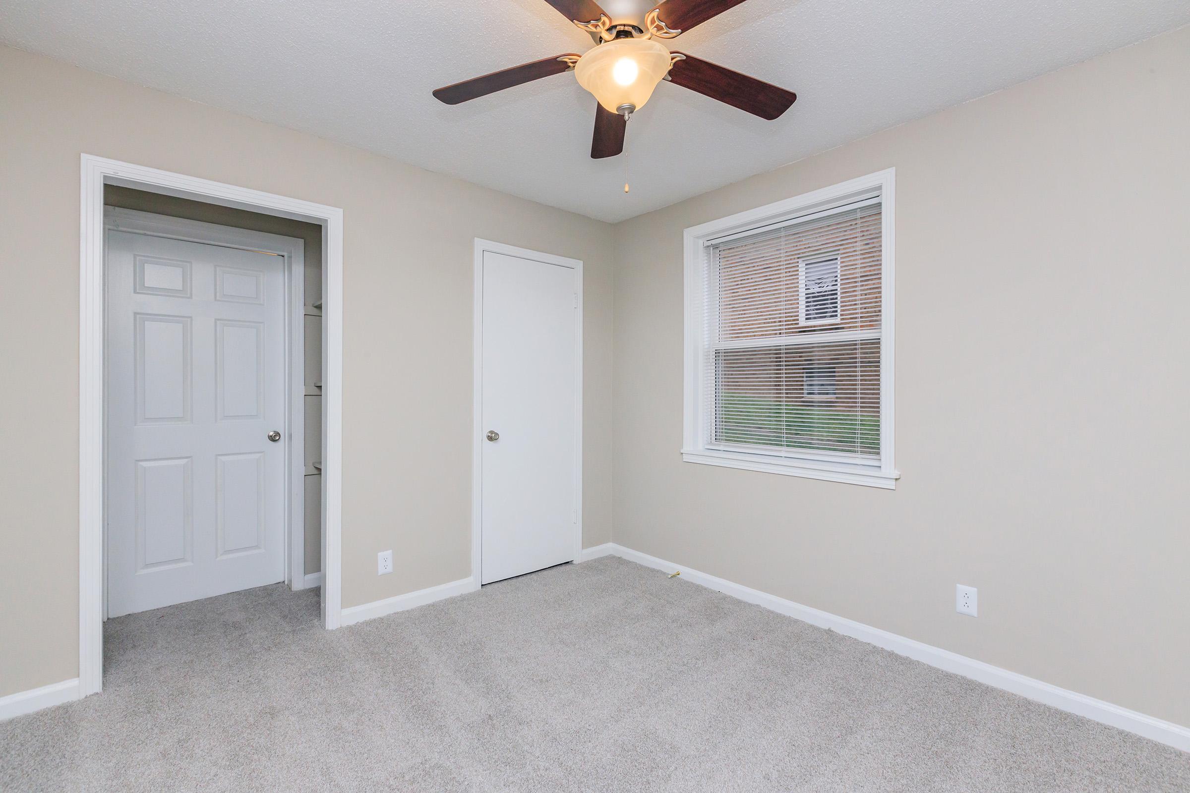 A well-lit, empty bedroom featuring light beige walls, a ceiling fan, and gray carpet. There is a closed white door on the left leading to a closet, another closed white door straight ahead, and a window on the right allowing natural light to enter. The aesthetic is clean and modern.