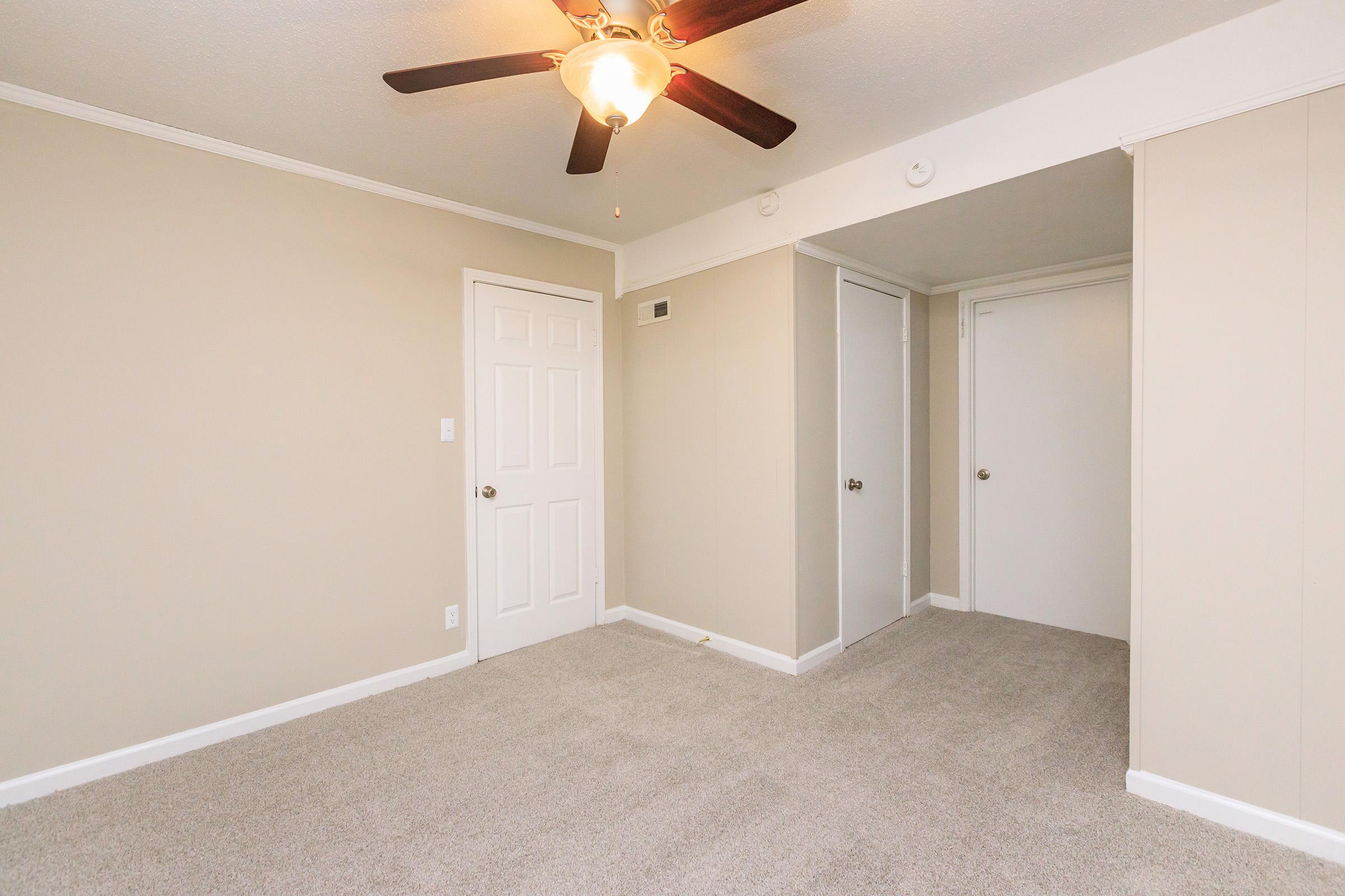 A clean, empty room with beige walls and light gray carpet. A ceiling fan is mounted above, and there are two closed closet doors on one side. A white door leads to another area. The lighting is bright and the space appears recently renovated.