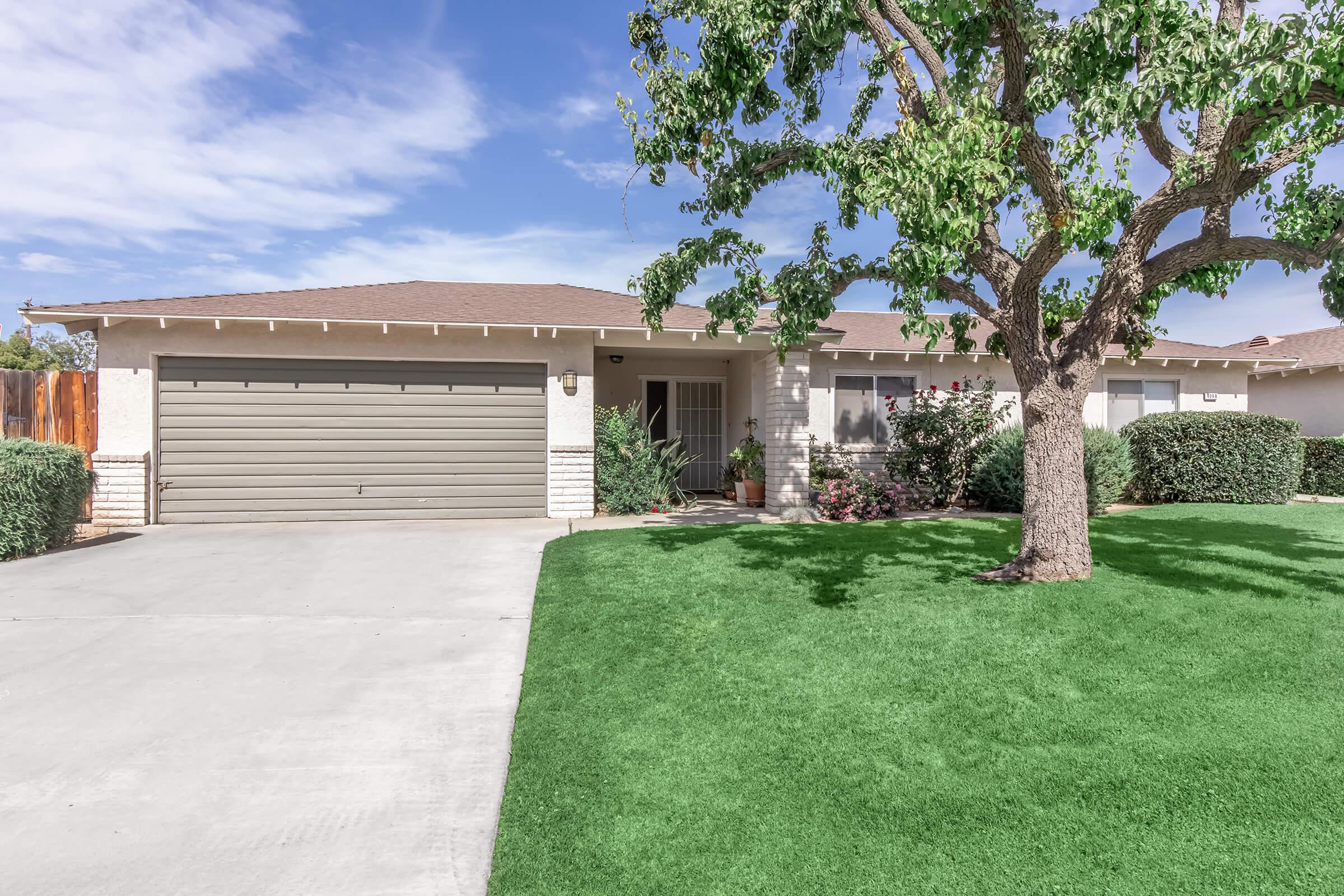 A single-story house with a light brown exterior and a gray garage door. The front yard features a well-manicured green lawn and a large tree. Flowering bushes are visible near the entrance, and the sky is blue with a few clouds.