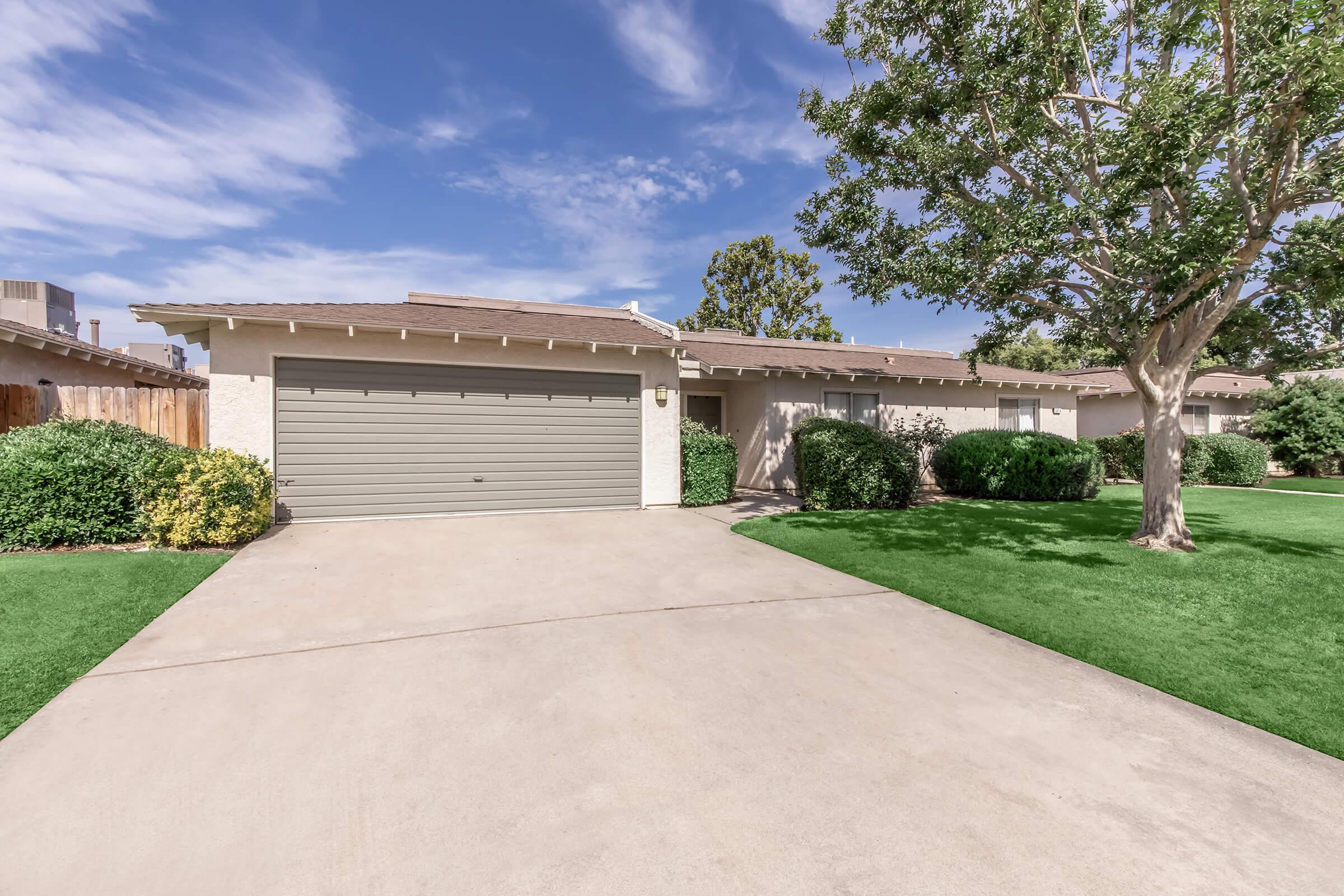 A single-story house with a gray garage door, surrounded by neatly trimmed bushes and a well-maintained lawn. The sky is partly cloudy, adding to the pleasant suburban atmosphere.
