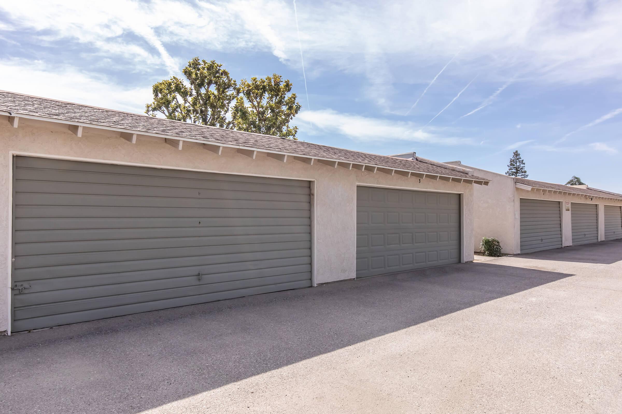 Row of garages with gray roll-up doors, situated along a paved driveway. The buildings are light-colored with a smooth finish, and there are trees in the background under a clear blue sky with some wispy clouds. The scene is well-lit and reflective of a suburban setting.