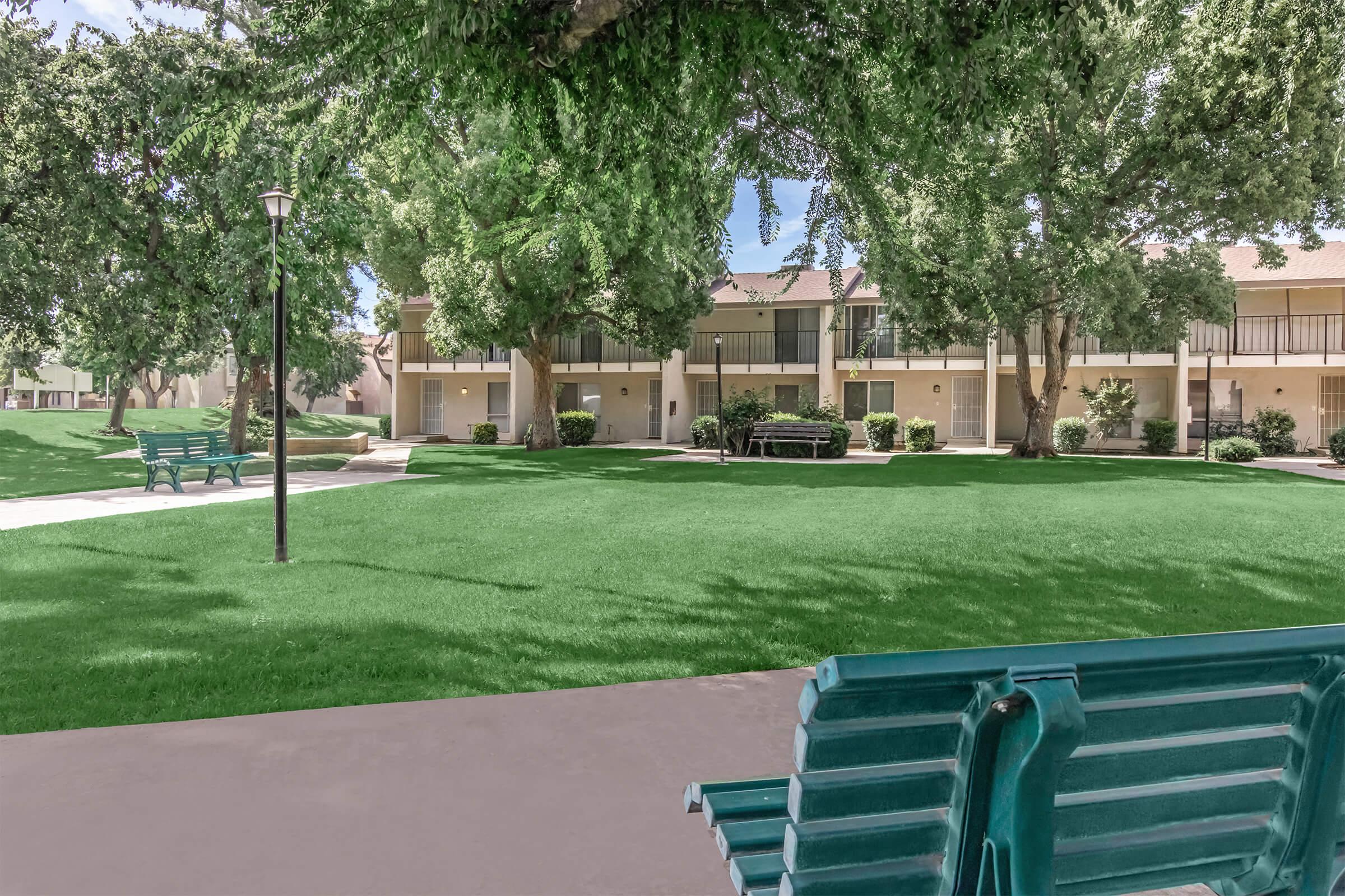 A green park area featuring well-maintained grass, shaded by large trees. In the foreground, there is a painted green bench. In the background, a row of apartment buildings can be seen, with a small gathering area that includes a grill and picnic tables. The setting is bright and inviting, ideal for relaxation.