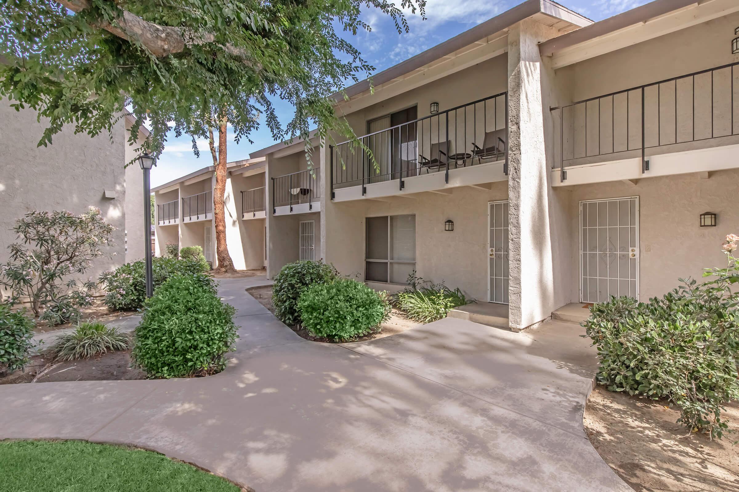 A tranquil exterior view of a residential complex featuring two-story buildings with balconies. Lush green shrubs line a concrete pathway, leading to individual entrances. Mature trees provide shade, and a clear blue sky is visible above, enhancing the peaceful atmosphere of the environment.