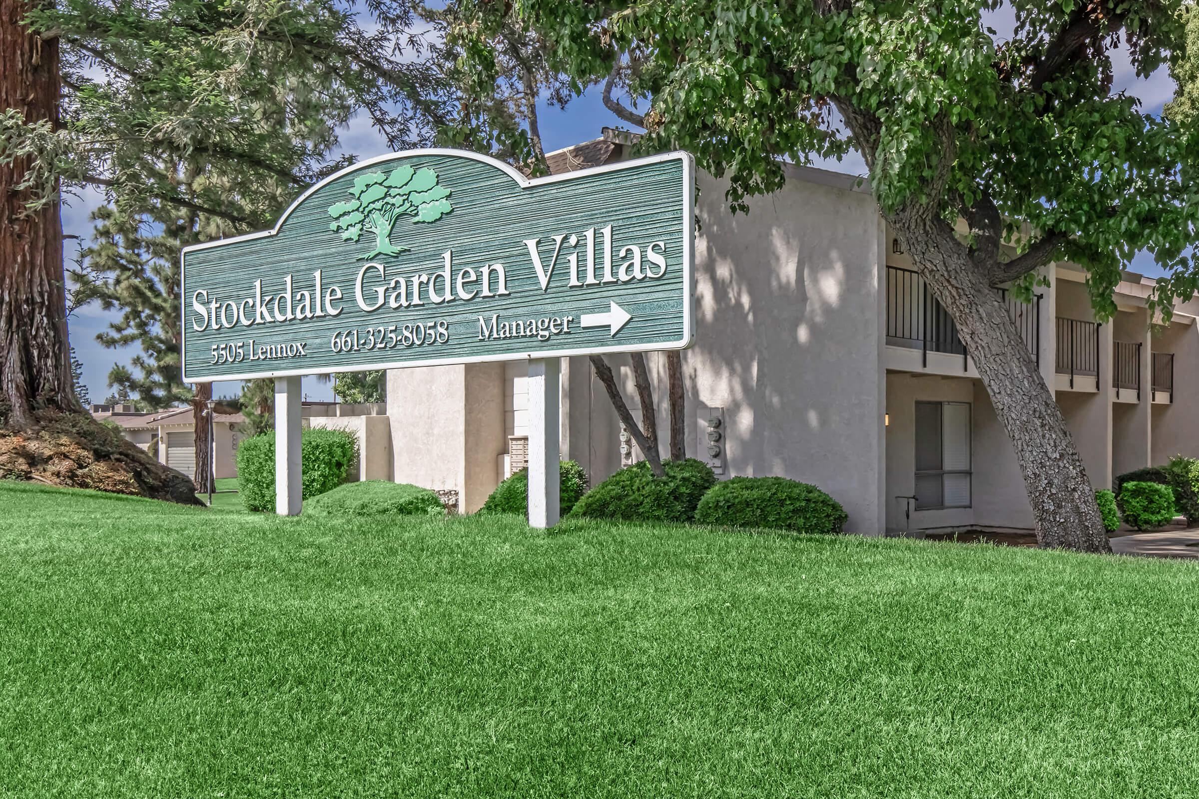 Sign for Stockdale Garden Villas with the address 3501 Lamoore, displaying the manager's contact information. Surrounding the sign is a grassy area and trees, with a clear sky in the background. The sign features a green color scheme and is set in a suburban residential environment.